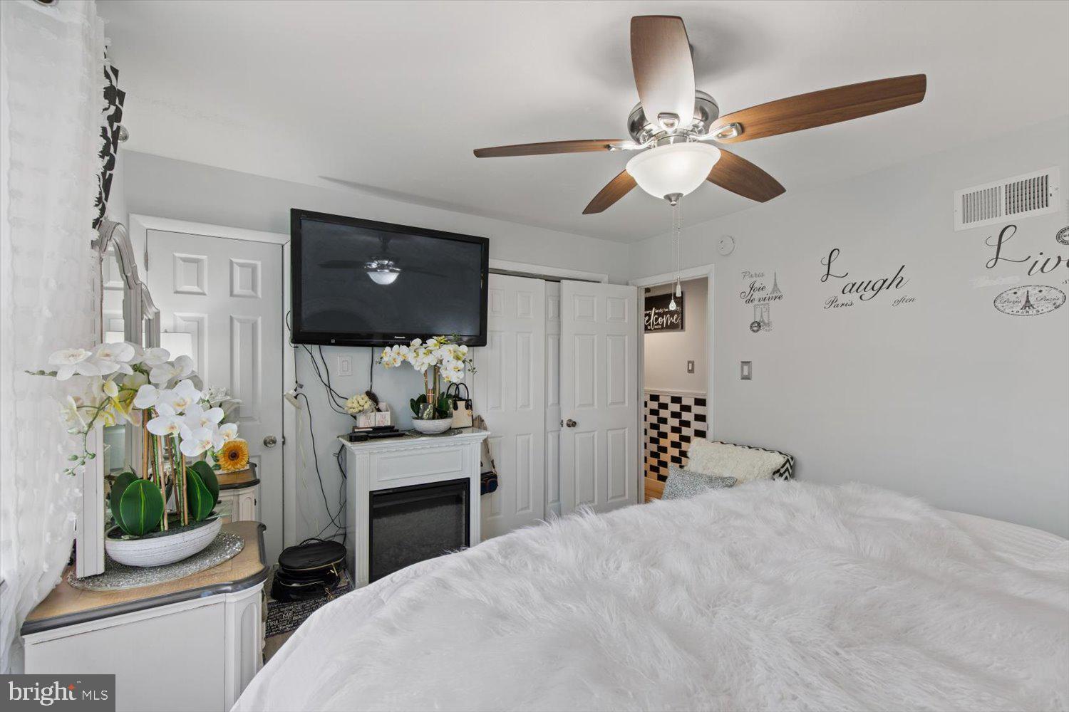 Bedroom with a white bed, wood-blade ceiling fan, and television above a fireplace.