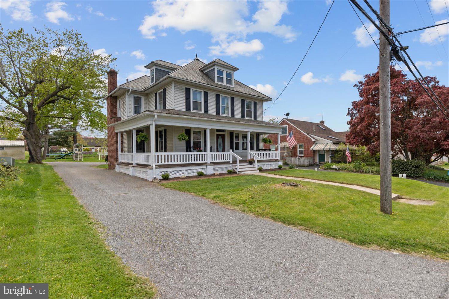 Two-story gray home with black shutters, a wraparound porch, dormers, and a gravel driveway.