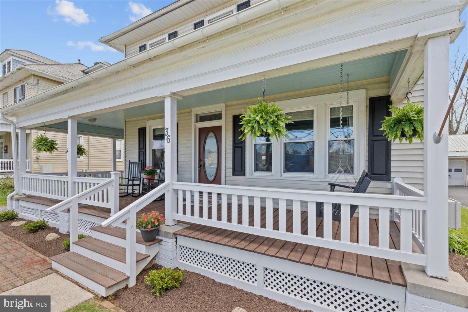 House front with a covered porch, white railings, brown decking, hanging ferns, and rocking chairs.