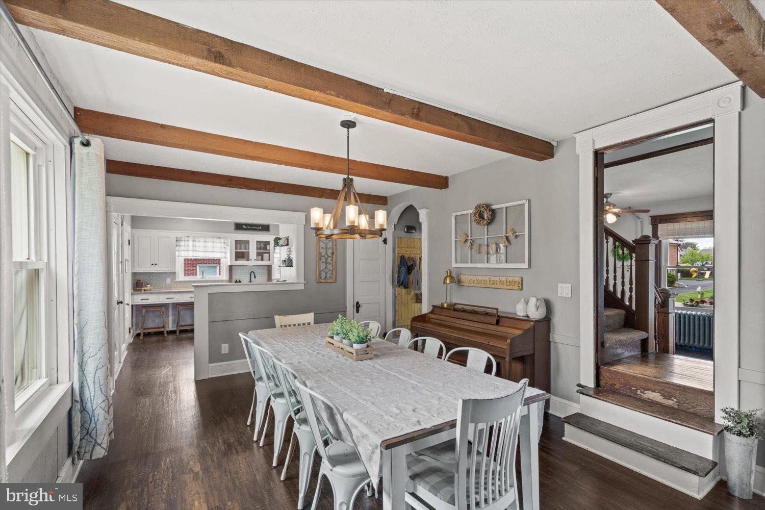 Dining room with exposed wood beams, a long table, and a piano beside the kitchen opening.