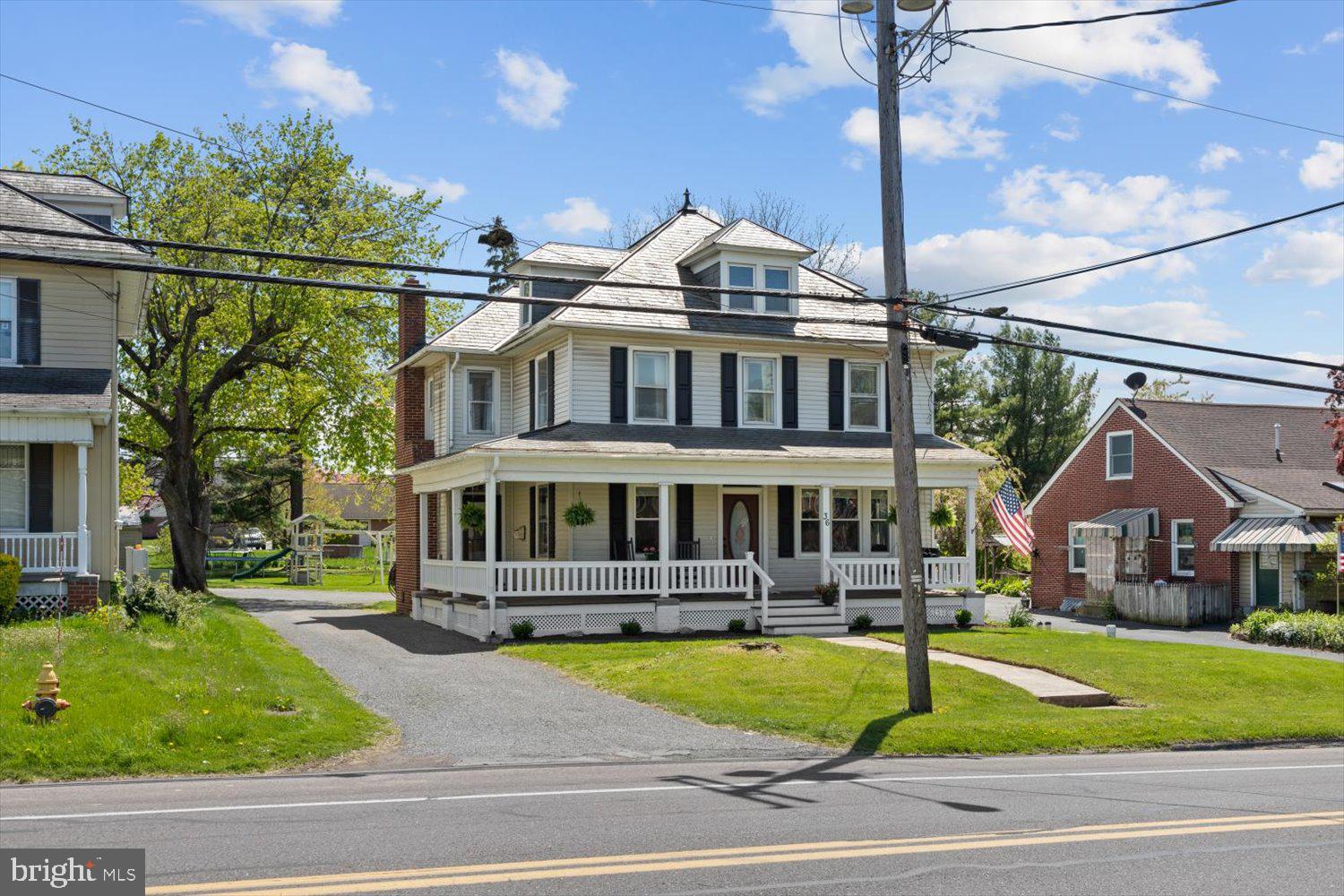 White two-and-a-half-story house with dark shutters and a wraparound porch beside a street.