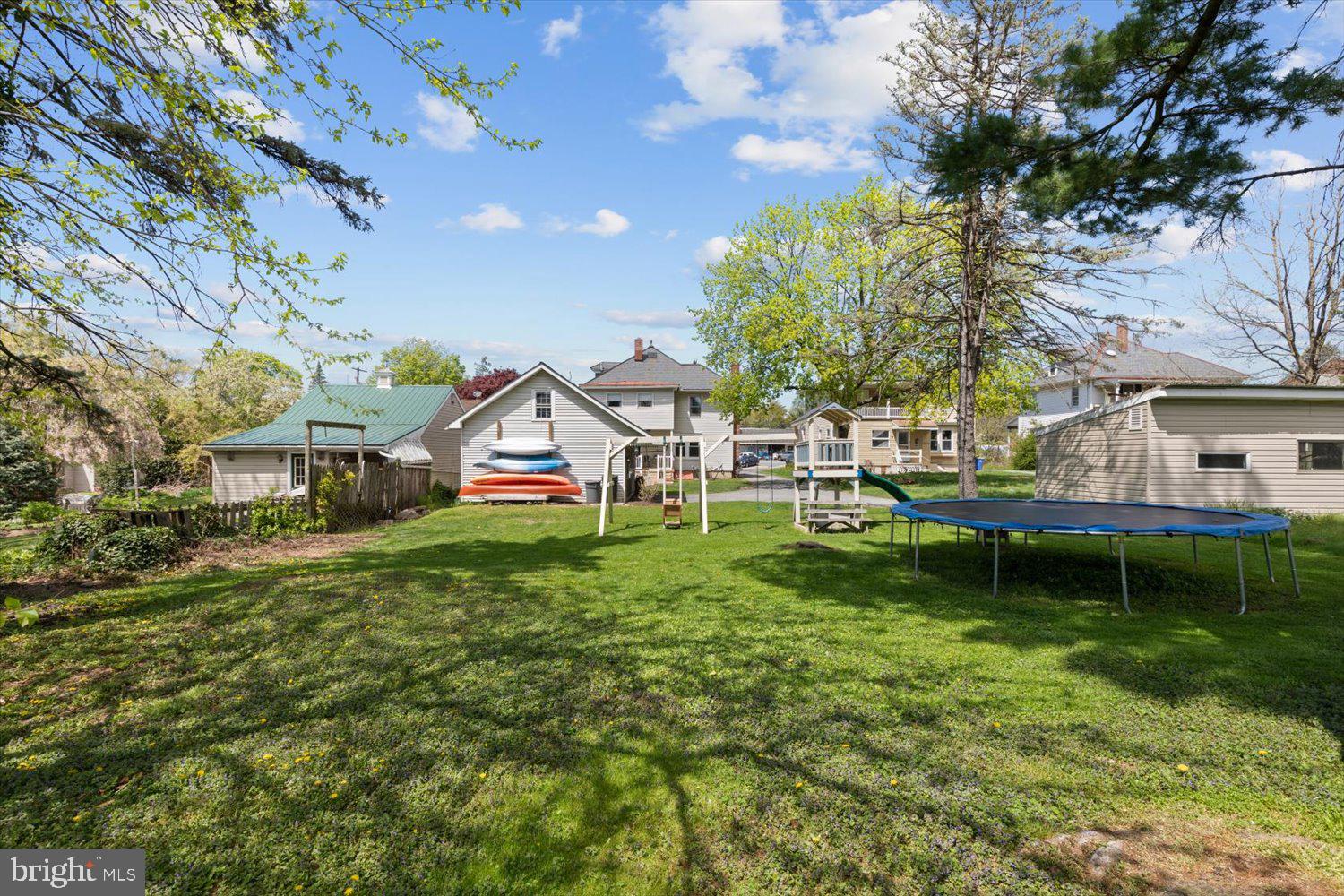 Grassy backyard with a trampoline, playset, kayaks, and outbuildings behind the house.