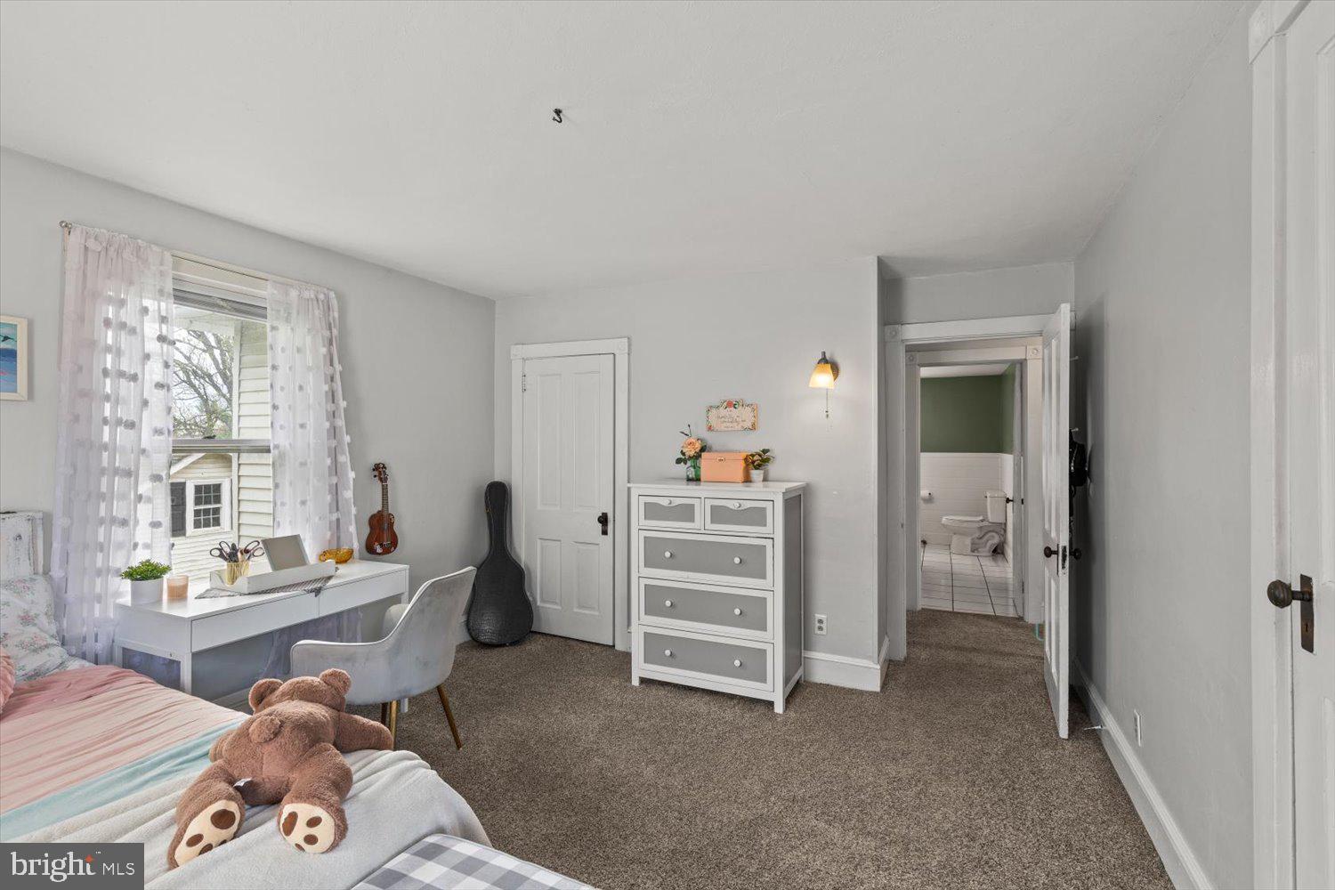 Bedroom with a teddy bear on the bed, a white desk by the window, and a bathroom doorway.