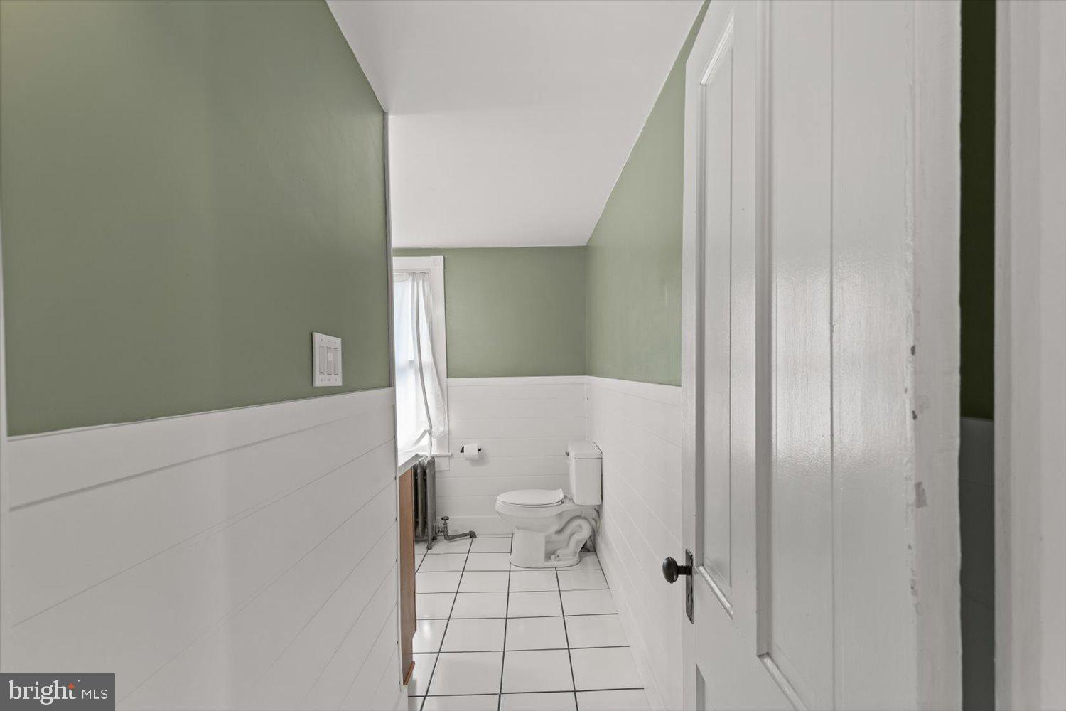 Narrow bathroom with sage-green walls, white tile, a toilet, and a window.
