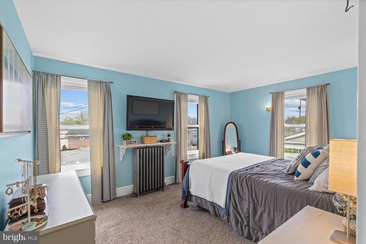 Bedroom with blue walls, a gray bed, three curtained windows, and a wall-mounted television.