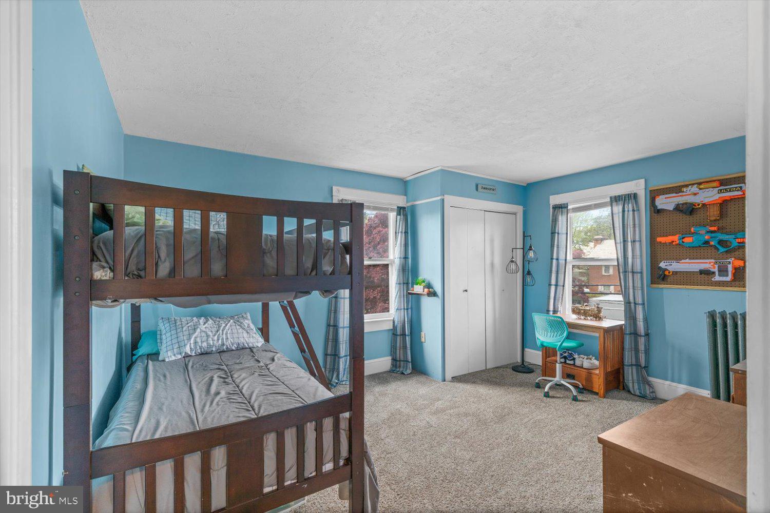 Blue bedroom with a dark wood bunk bed, two curtained windows, and a desk beneath a toy blaster display.