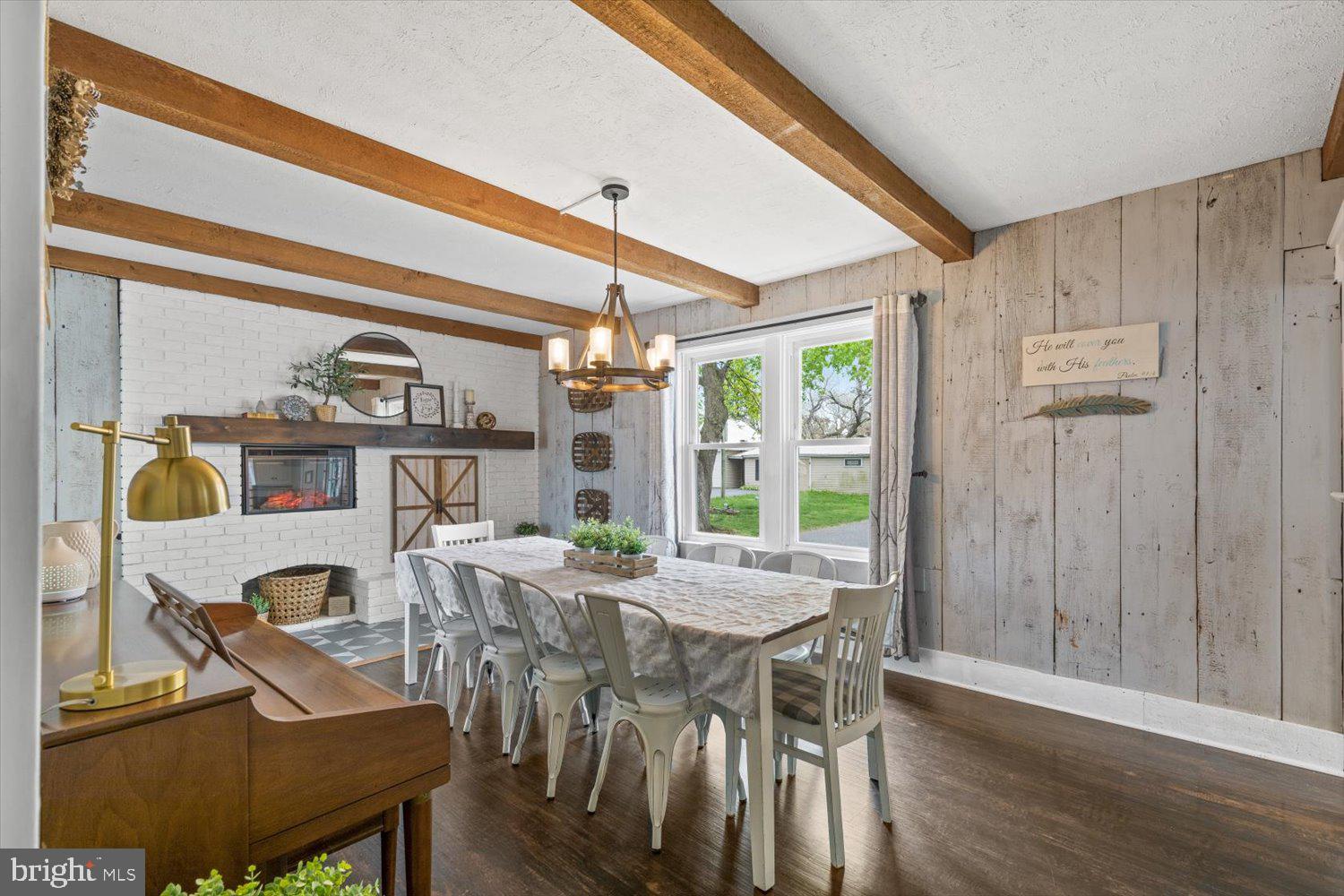 Dining room with exposed wood beams, a white brick fireplace, and a long table by large windows.