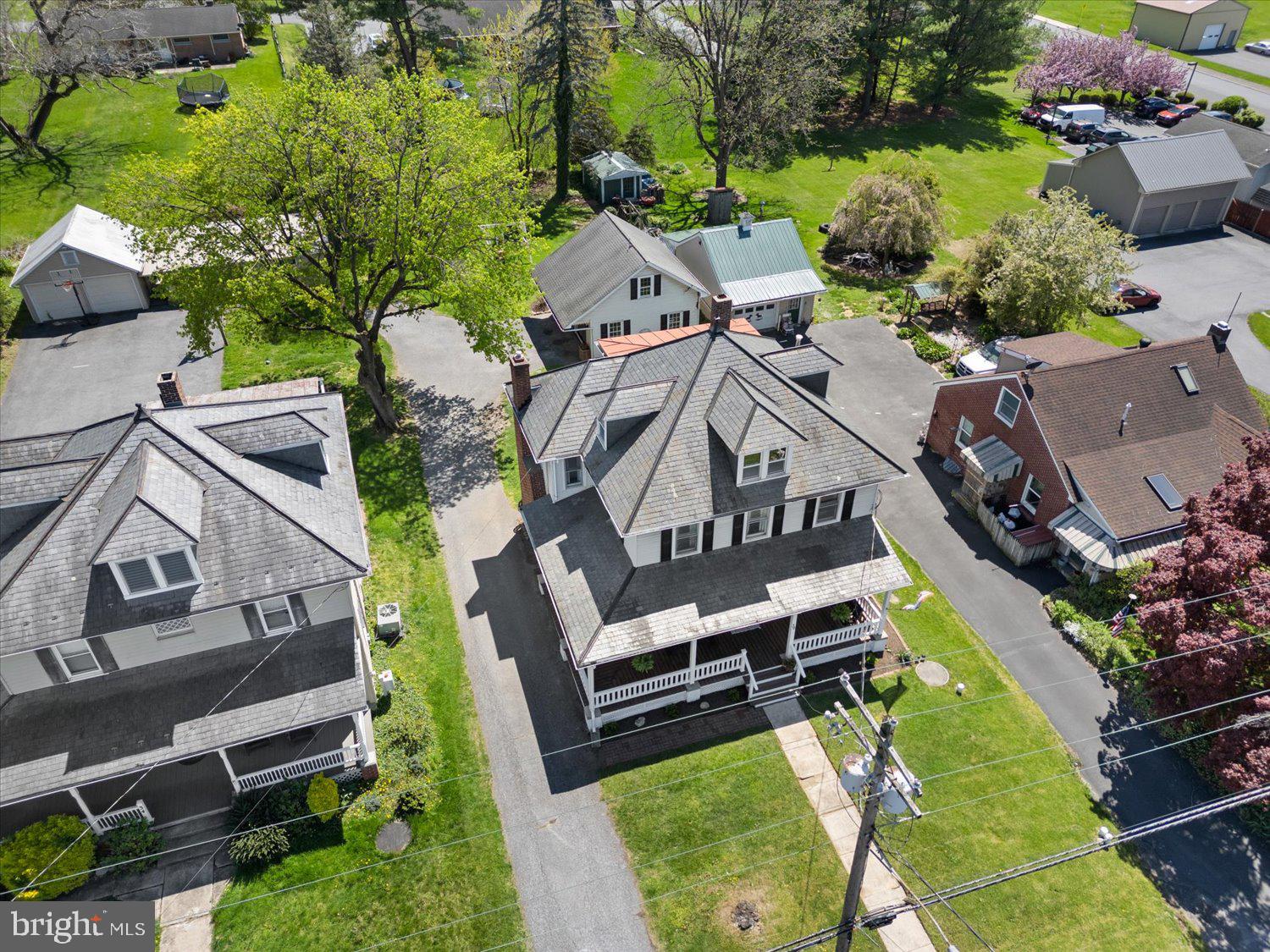 Aerial view of a house with a broad gray shingle roof, covered porch, paved driveway, and mature trees.
