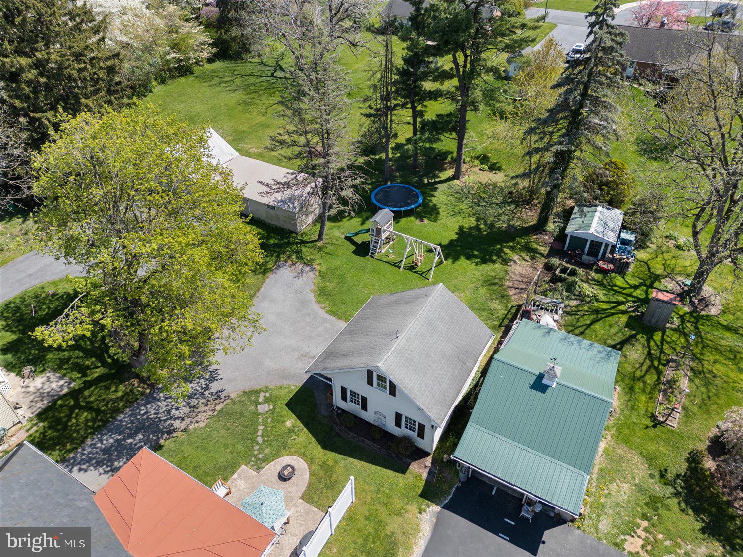 Aerial view of a tree-lined backyard with a trampoline, playset, outbuildings, and paved areas.