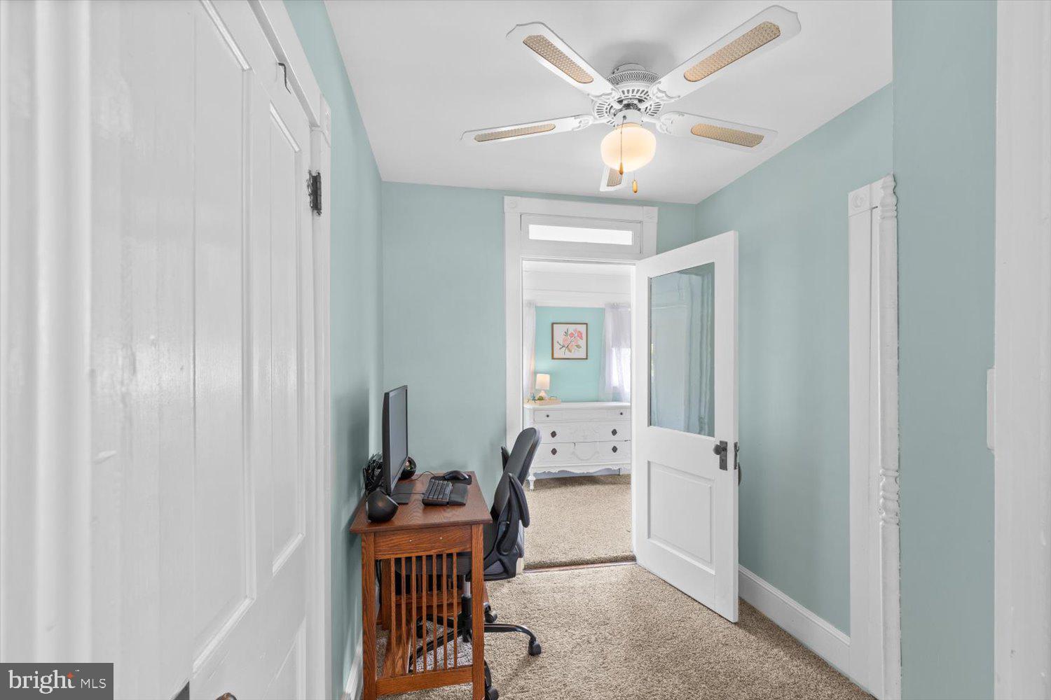 Home office with a wooden computer desk, pale aqua walls, and an open doorway to a bedroom.