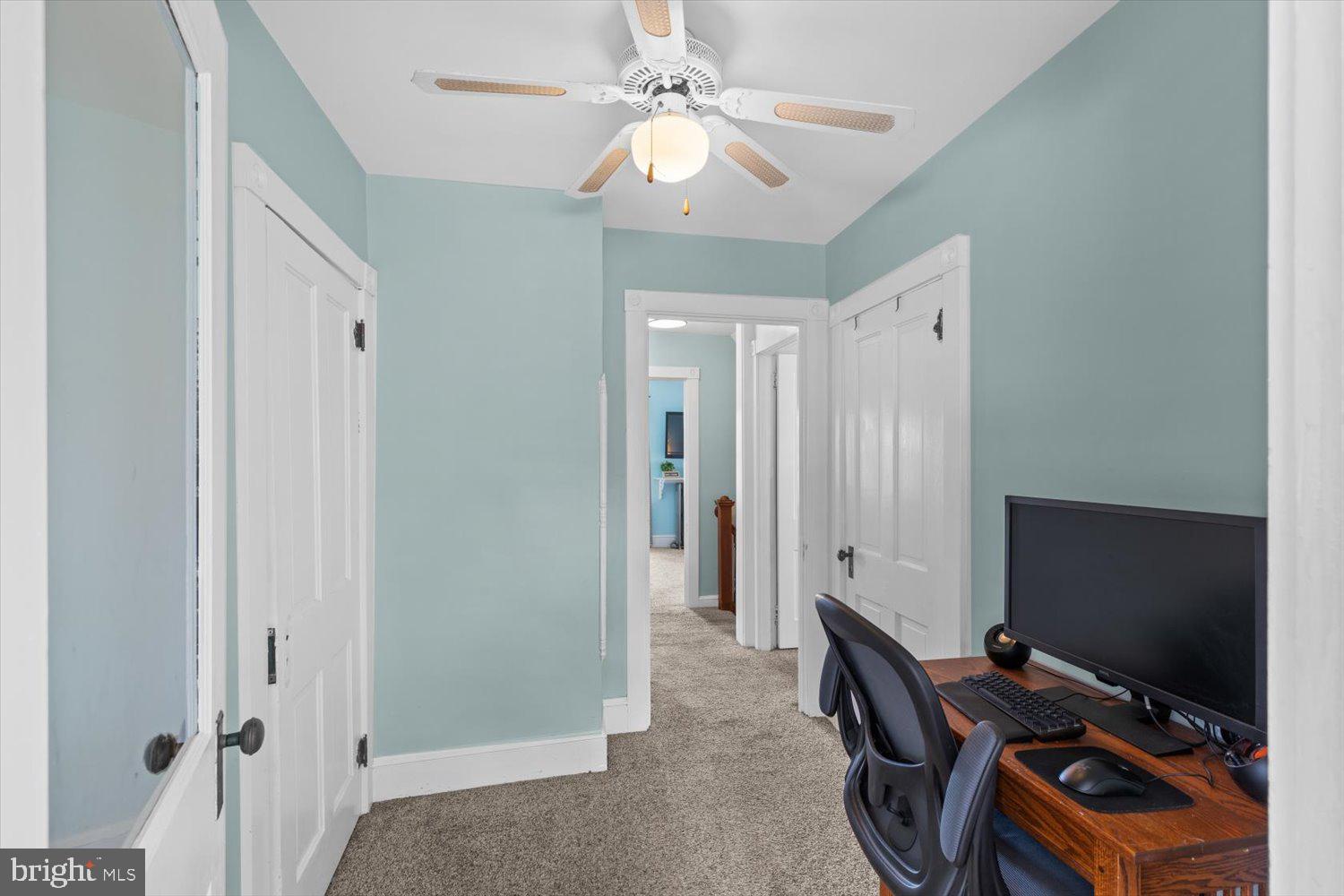 Home office with a wooden computer desk, pale blue-green walls, carpet, and a ceiling fan.