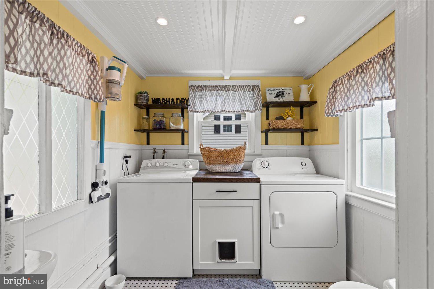 Laundry room with white washer and dryer, yellow walls, patterned curtains, and open shelves.