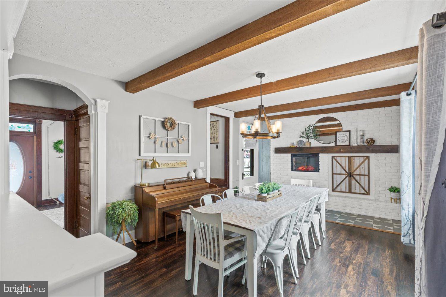 Dining room with exposed wood beams, a long table, piano, and white brick fireplace.