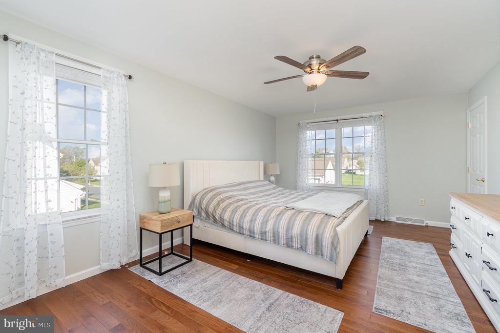 Bright bedroom with a large upholstered bed, hardwood floors, two curtained windows, and a ceiling fan.