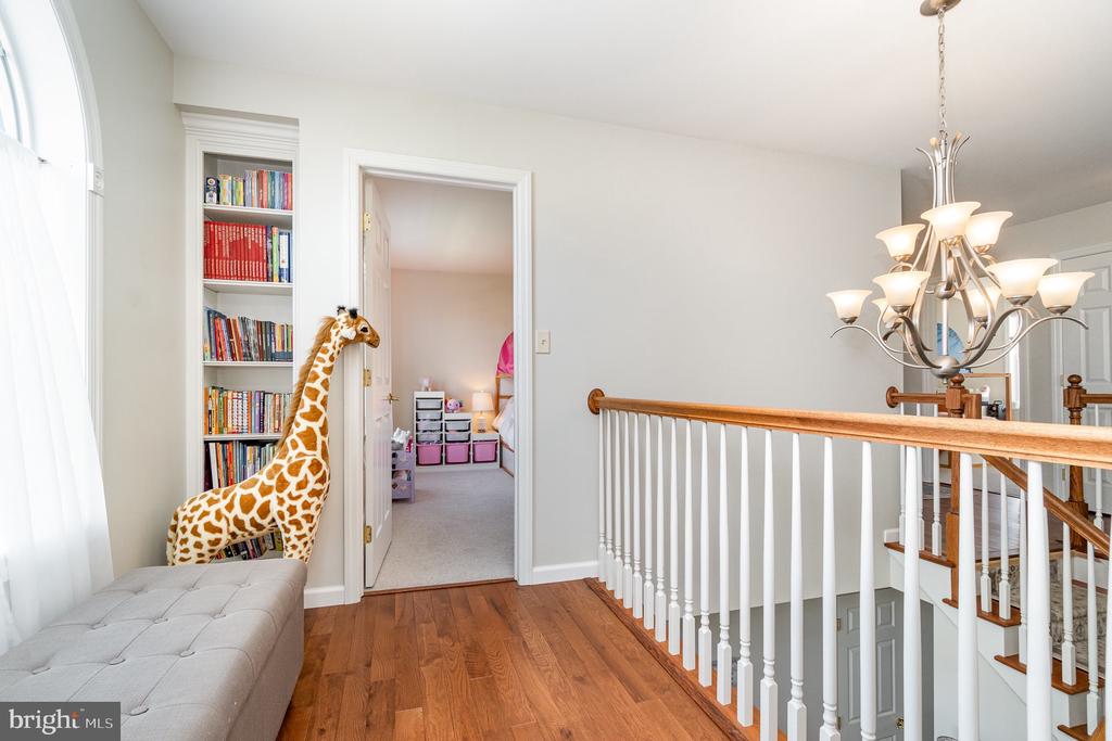 Upstairs hallway with hardwood floors, white stair railing, chandelier, and a giraffe toy beside a bookshelf.