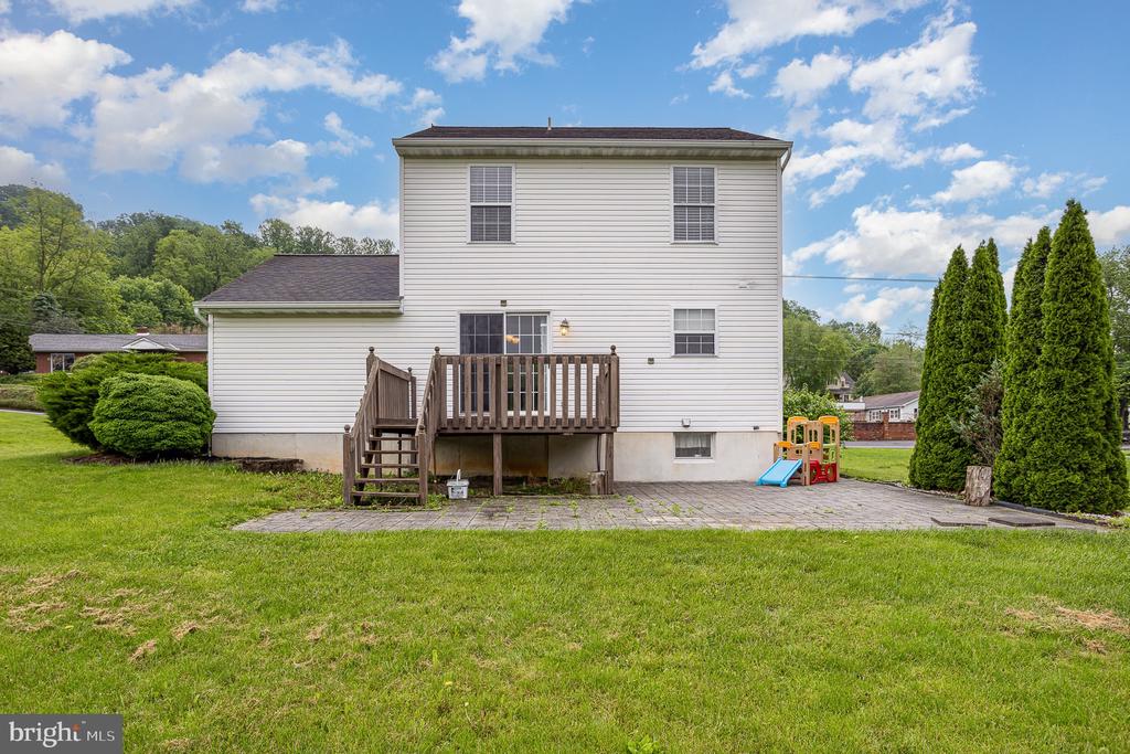Rear of a two-story white house with a raised deck, paved patio, lawn, and children’s play equipment.