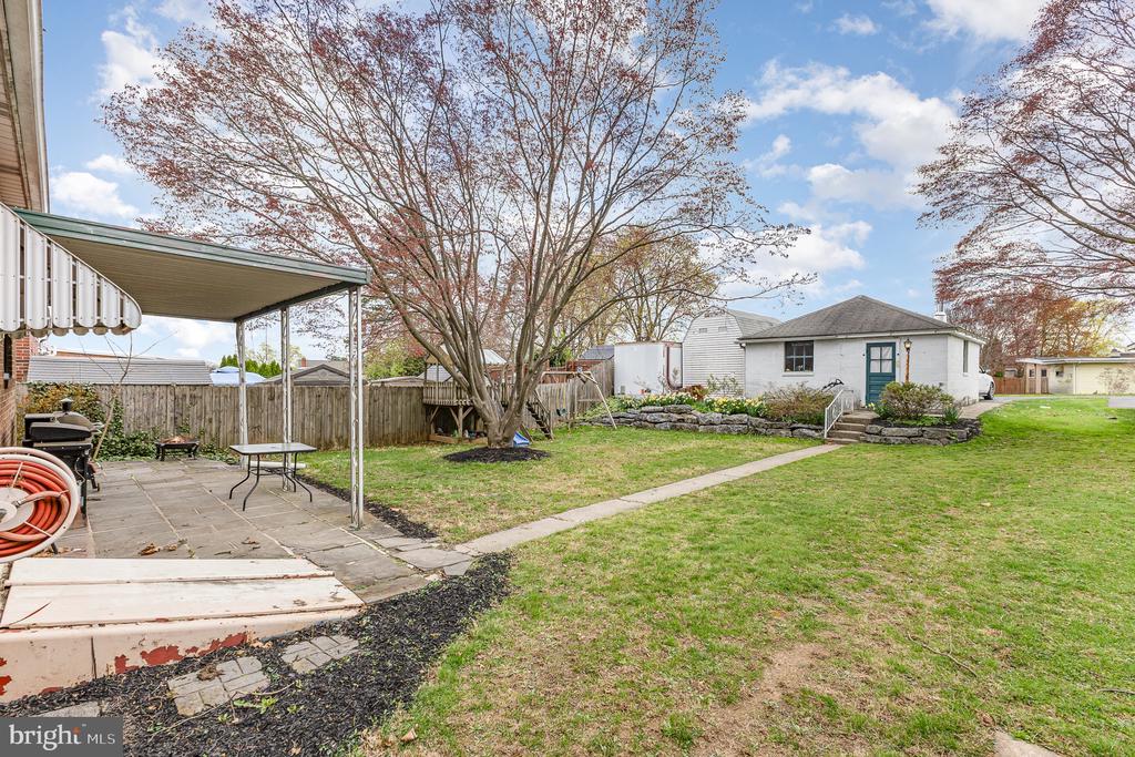 Backyard with a covered patio, large budding tree, lawn, and white outbuilding with a teal door.