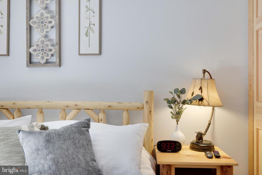 Bedroom with a natural wood bed frame, gray pillows, and a bedside table with a lamp and plant.