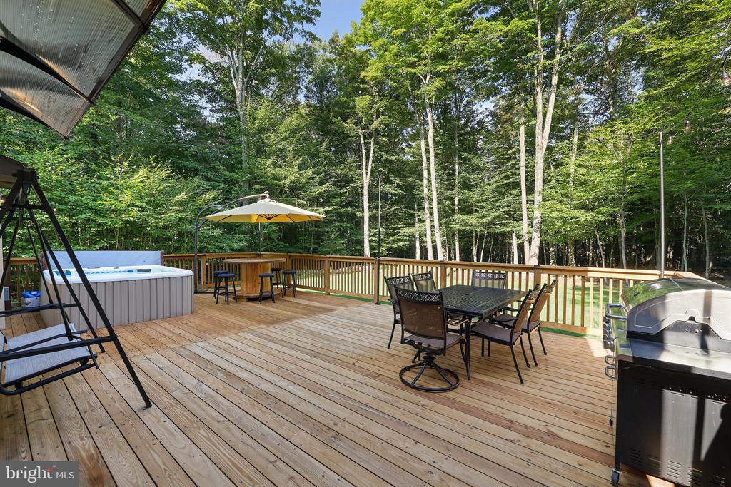 Wooden deck with a hot tub, outdoor dining set, grill, and surrounding woods.
