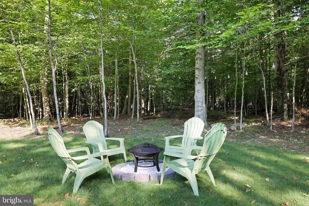 Backyard fire pit surrounded by pale green Adirondack chairs, with woods beyond.