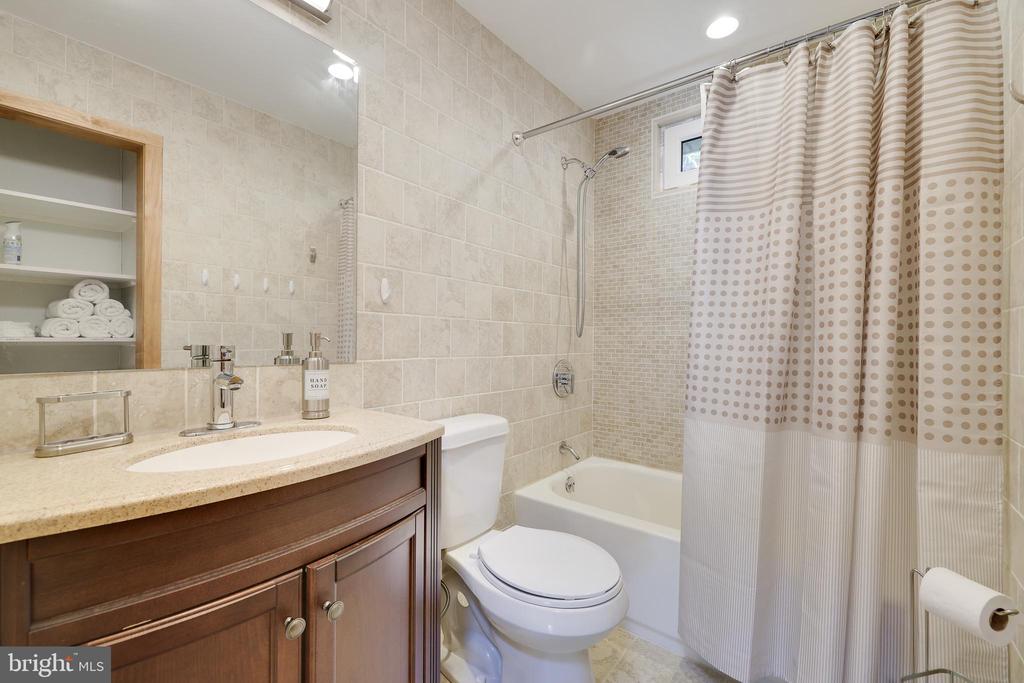 Bathroom with a wood vanity, tiled tub surround, patterned shower curtain, and recessed towel shelf.