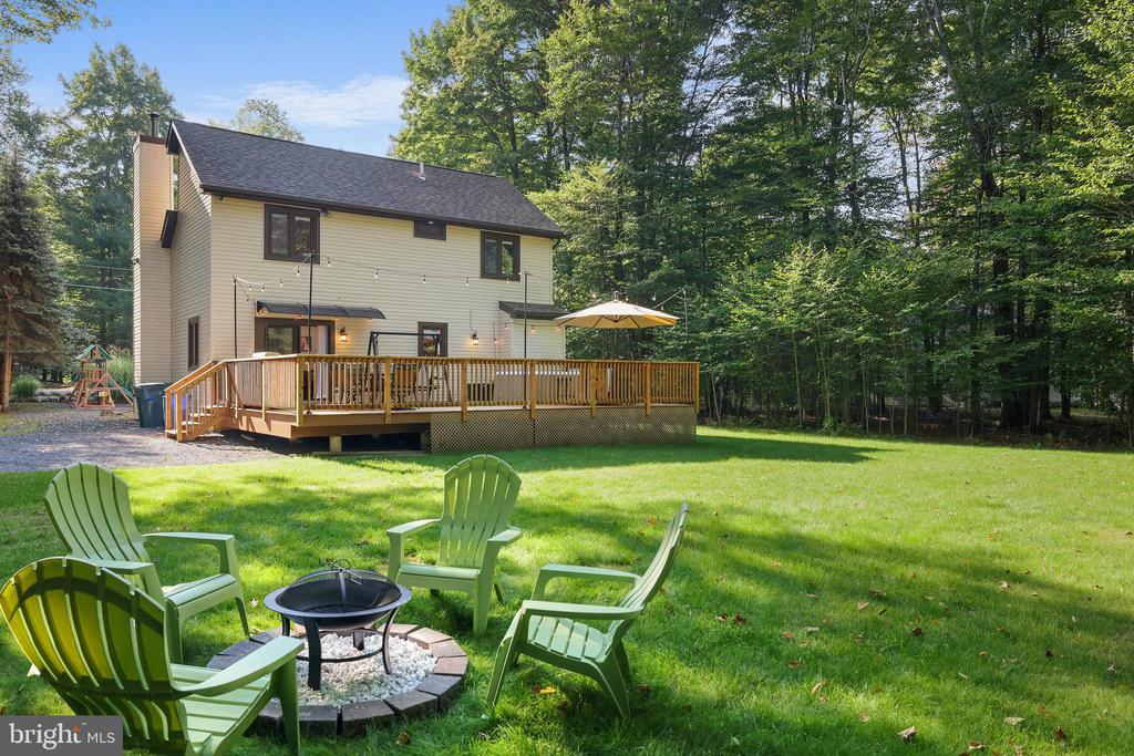 Two-story house with a large deck, lawn, Adirondack chairs, and a fire pit beside the woods.