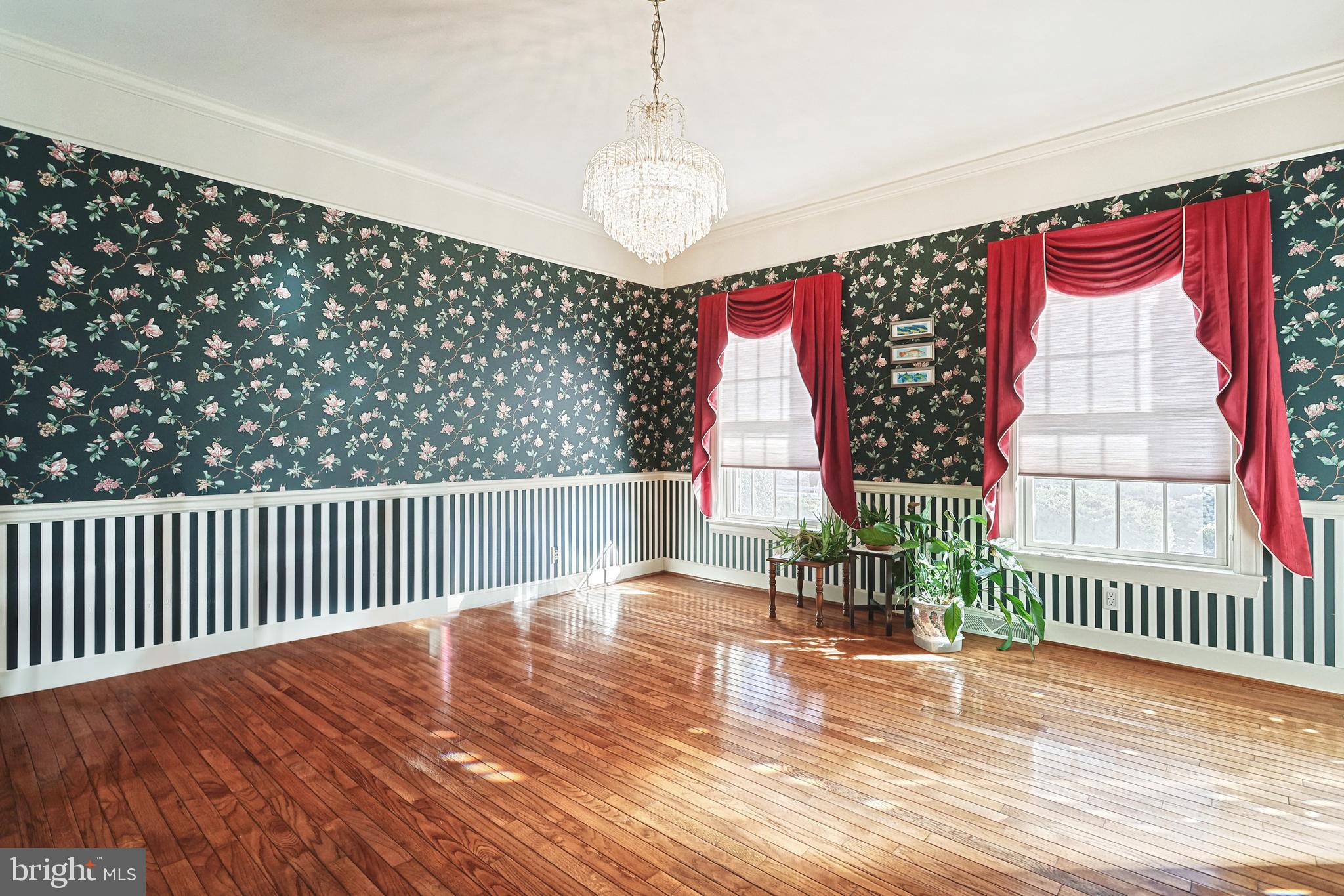Room with floral wallpaper, striped wainscoting, red curtains, polished hardwood floors, and a crystal chandelier.