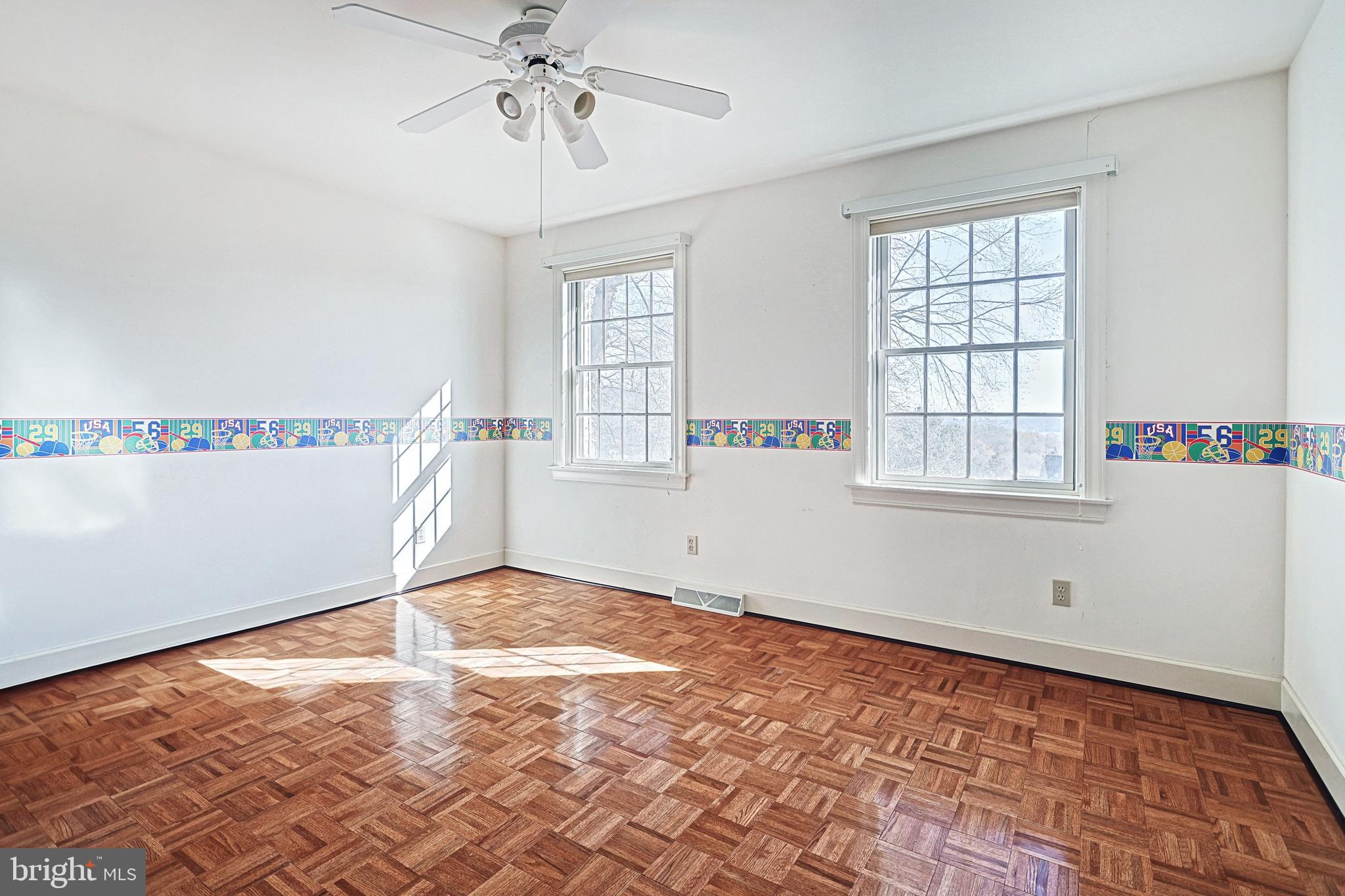 Vacant bedroom with parquet flooring, two tall windows, and a colorful sports-themed wall border.