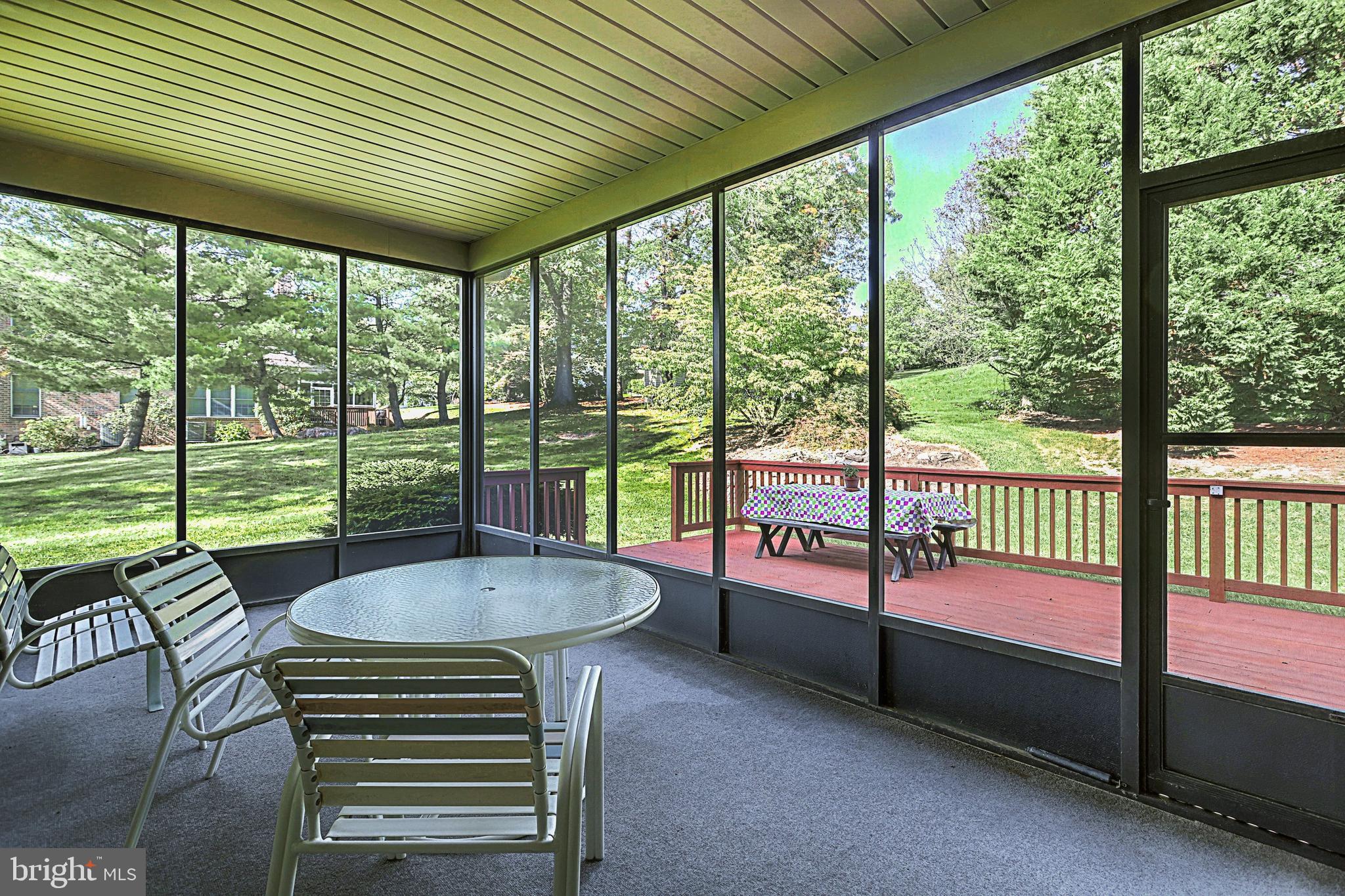 Furnished sunroom with large windows overlooking a deck and leafy yard.