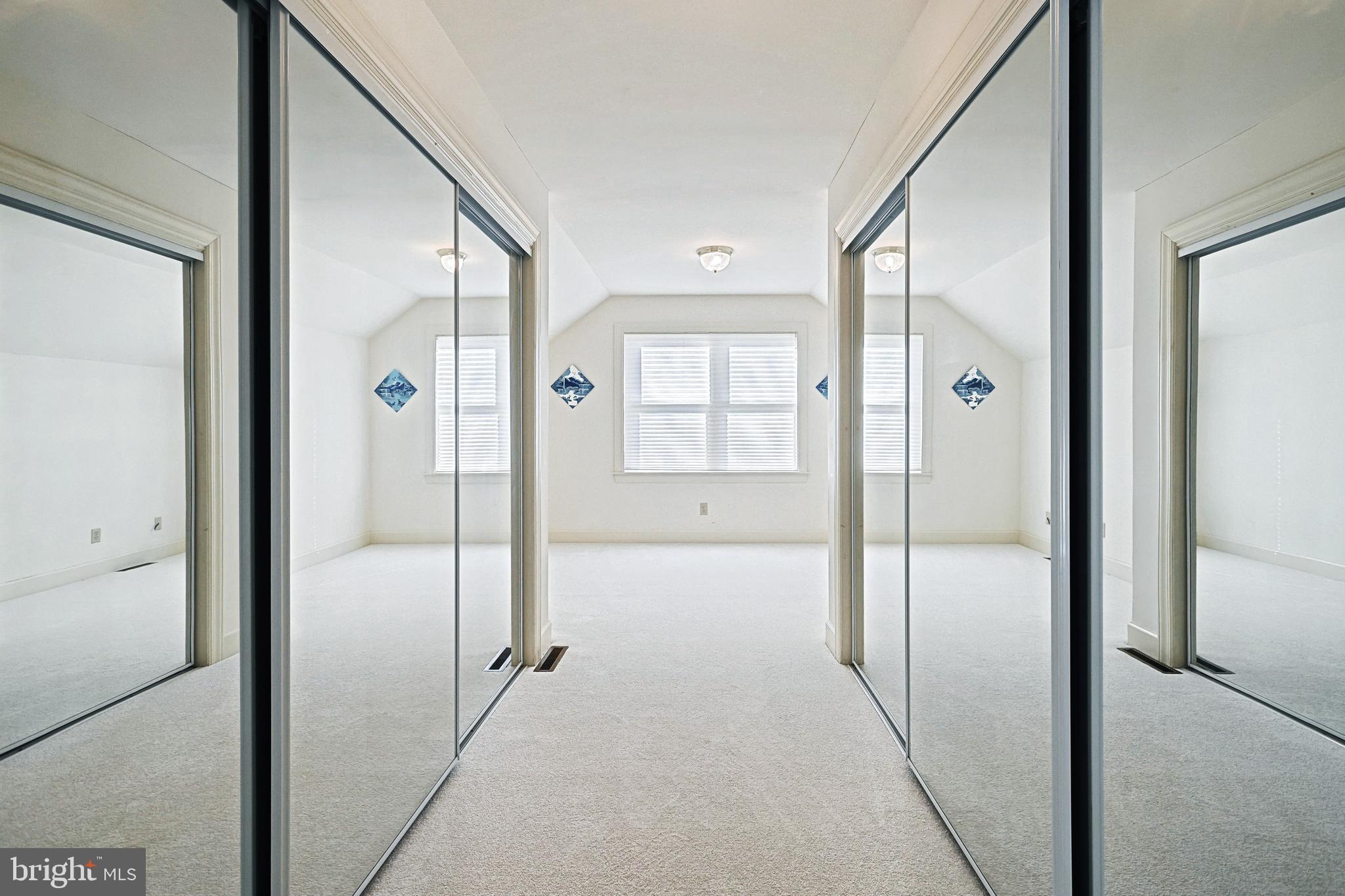 Empty carpeted bedroom with mirrored closet doors and windows with blinds.