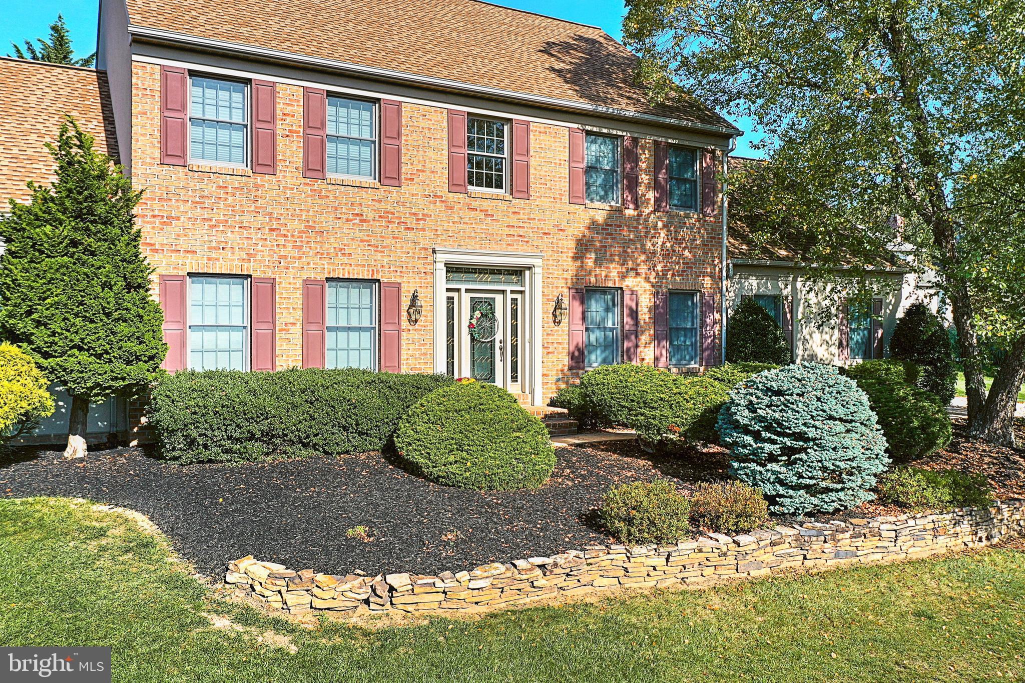 Two-story brick home with red shutters, landscaped shrubs, and a curved stone retaining wall.