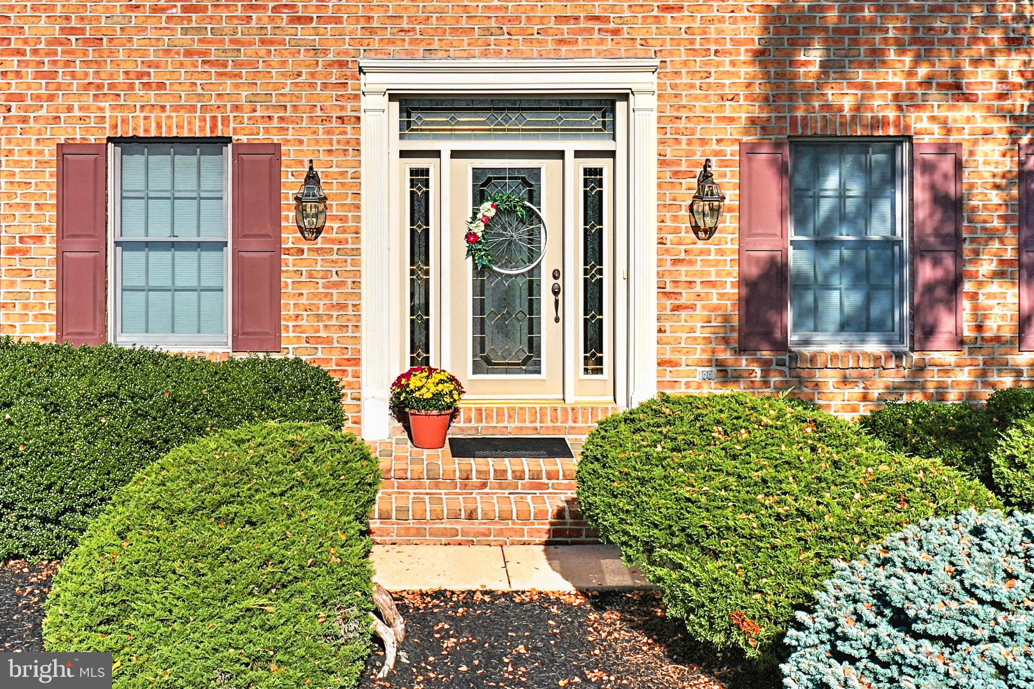 Brick home entrance with decorative glass door, wreath, brick steps, and trimmed shrubs.