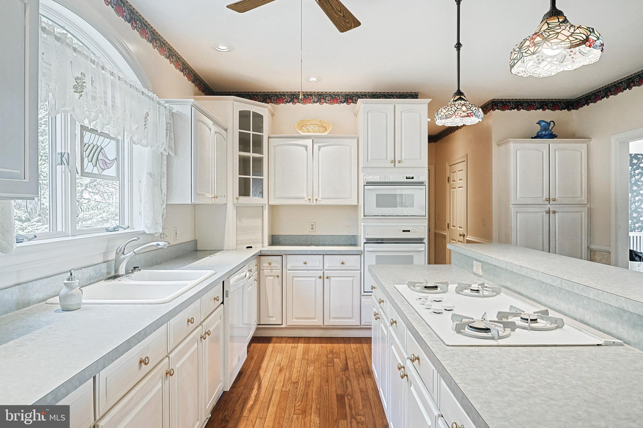 Bright kitchen with white cabinetry, island gas cooktop, double wall ovens, and stained-glass pendant lights.