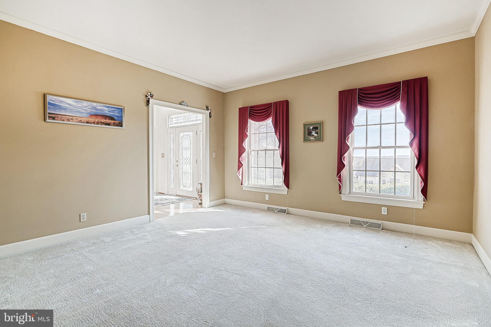 Empty carpeted living room with tan walls, burgundy window curtains, and a decorative glass-paneled entry door.