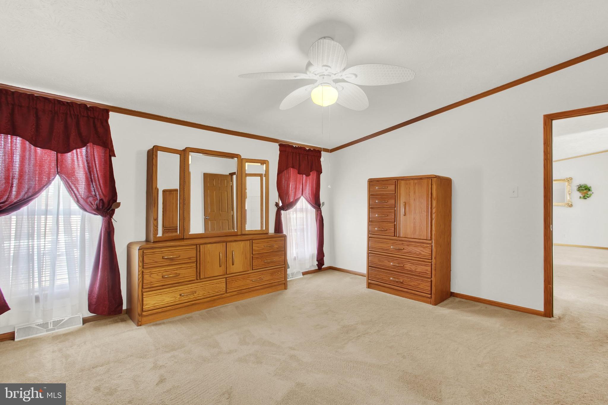 Furnished bedroom with wooden dressers, red curtains, beige carpet, and a ceiling fan.
