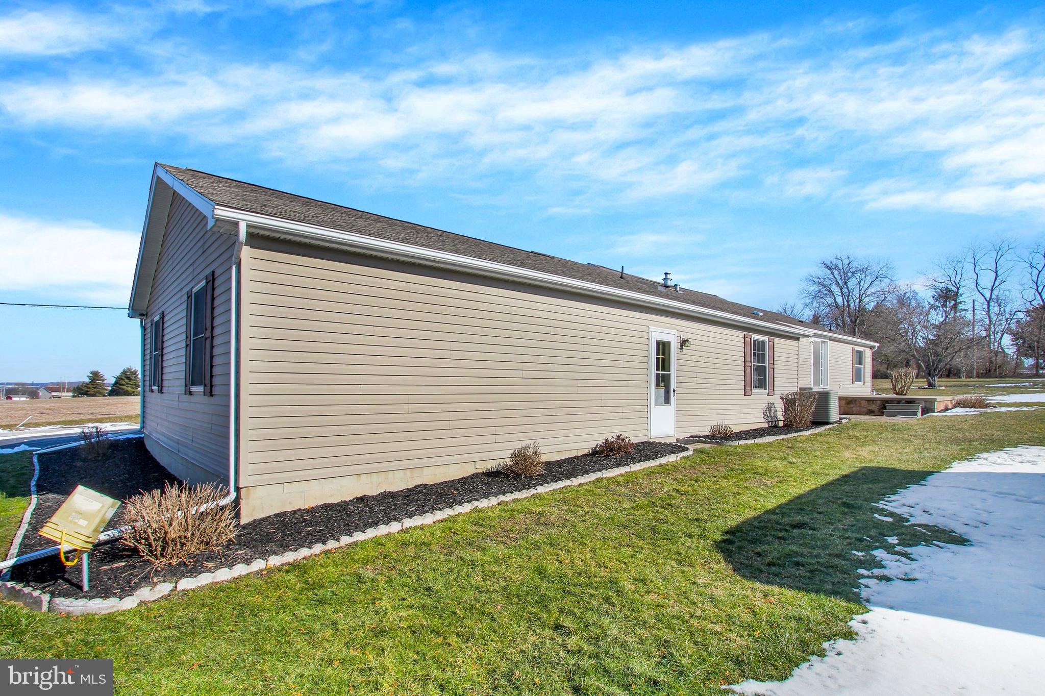 Side of a beige-sided home with a long roofline, foundation beds, and patches of snow in the yard.