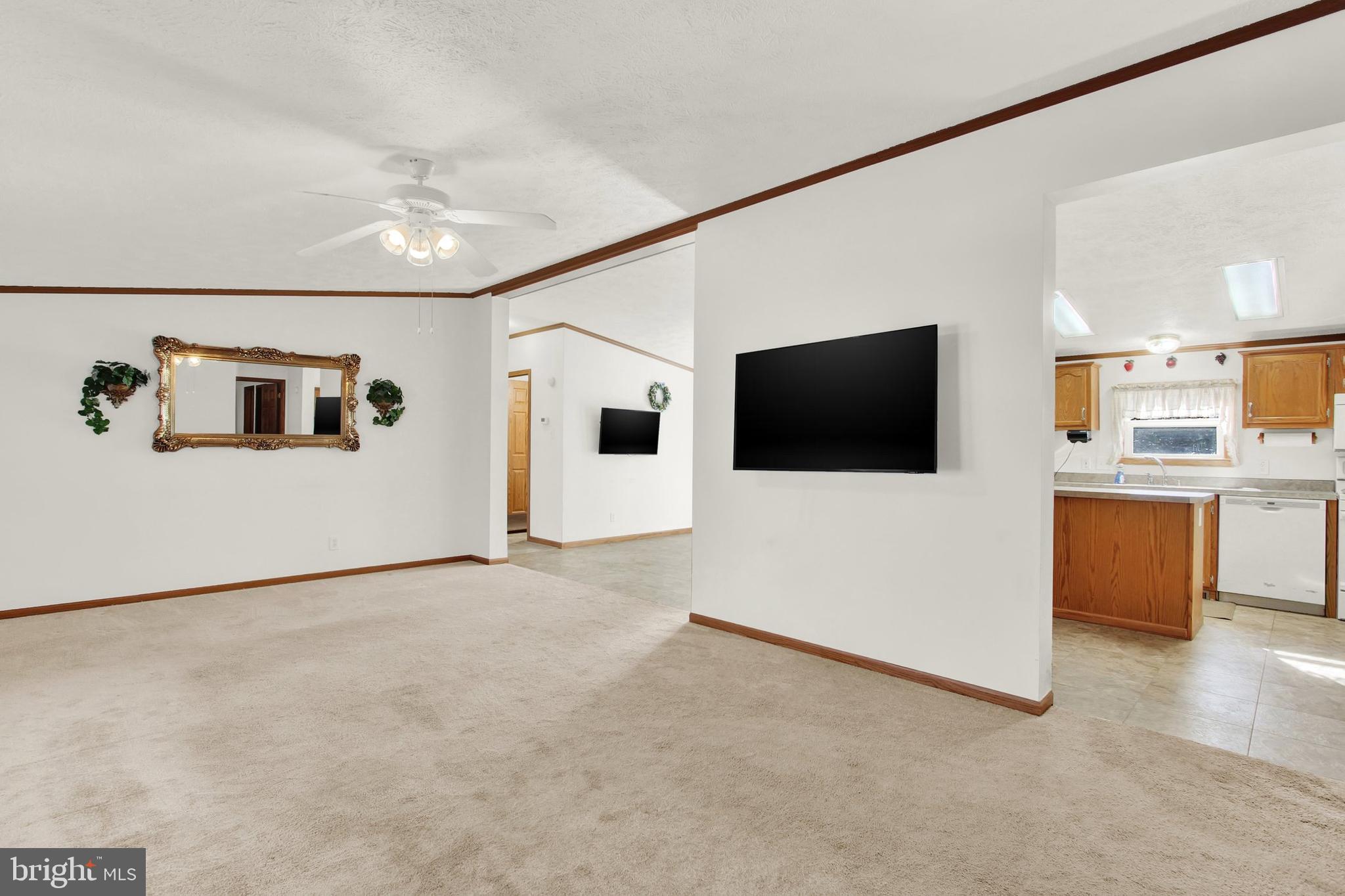 Living room with beige carpeting and ceiling fan, opening onto a kitchen with wood cabinets.