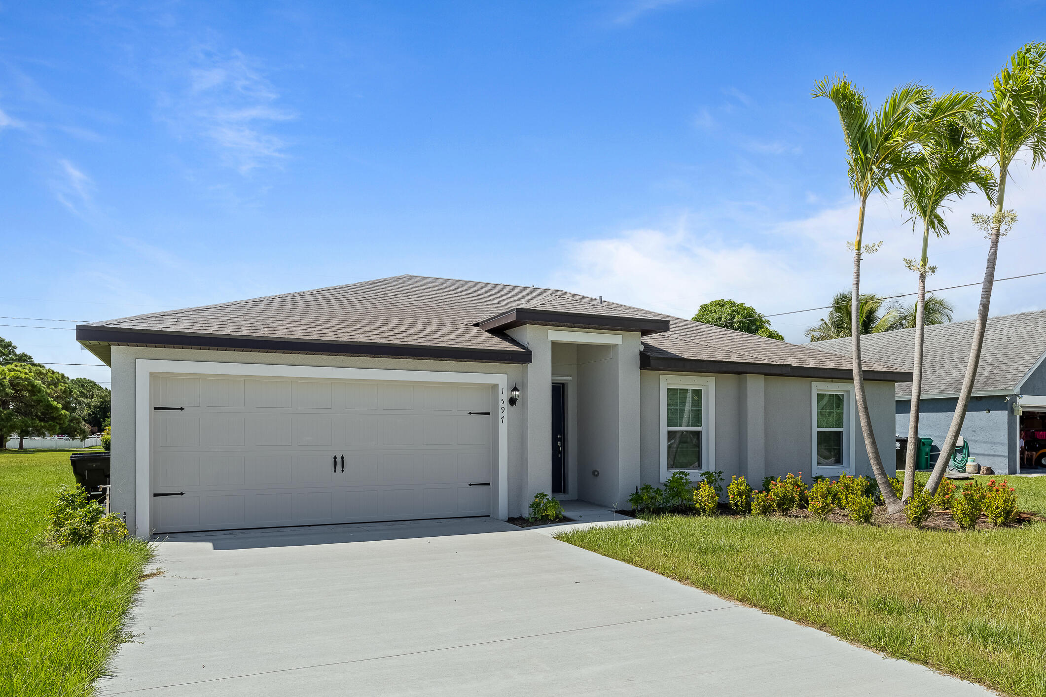 Single-story gray home with a wide garage, landscaped lawn, and tall palm trees.