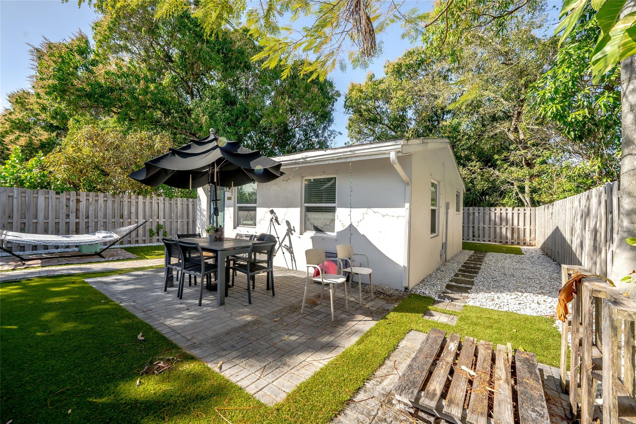 Backyard patio with outdoor dining set, black umbrella, hammock, and fenced gravel walkway.