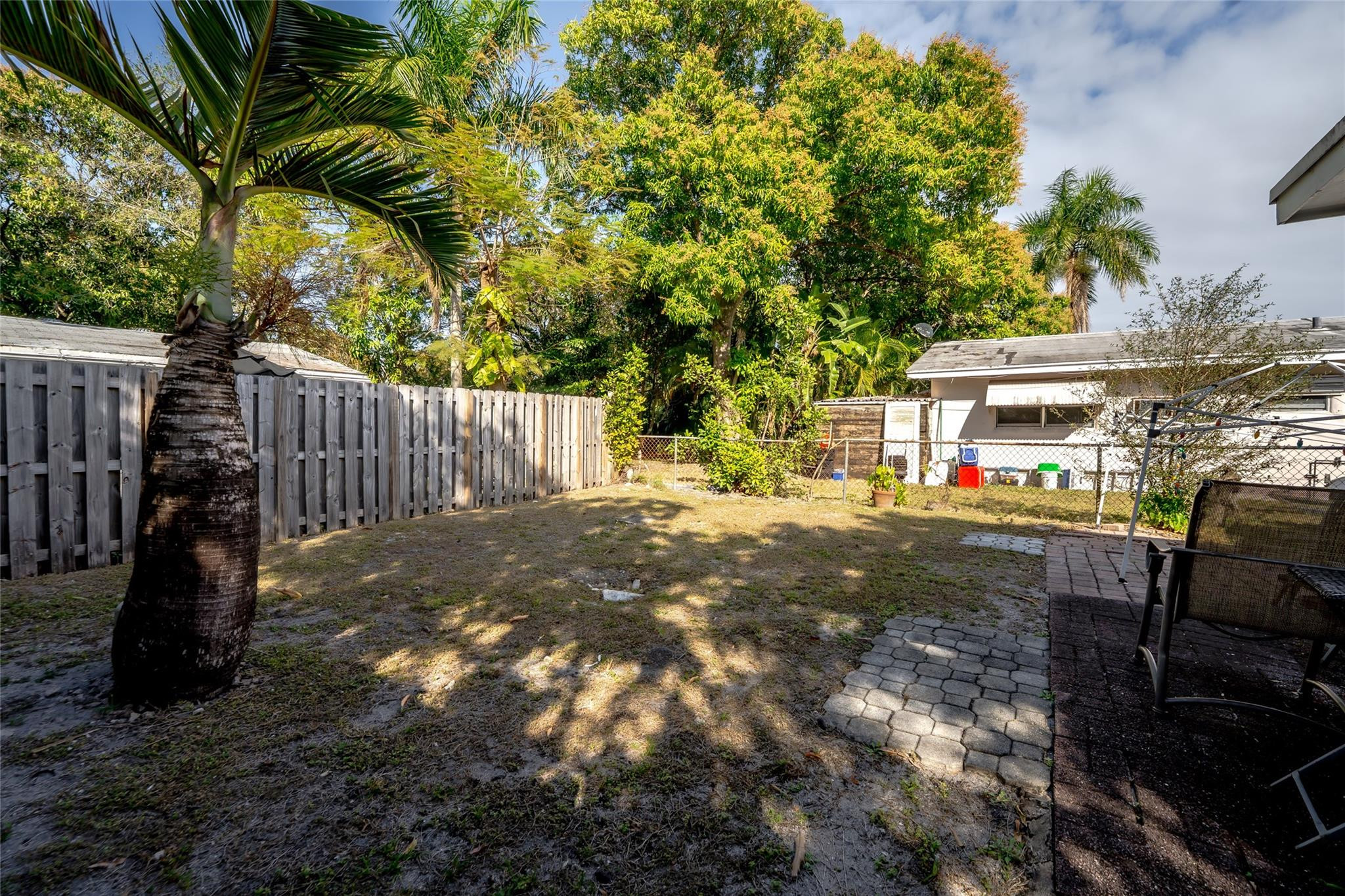 Backyard with mature palm trees, a wooden privacy fence, and a paved patio with chairs.