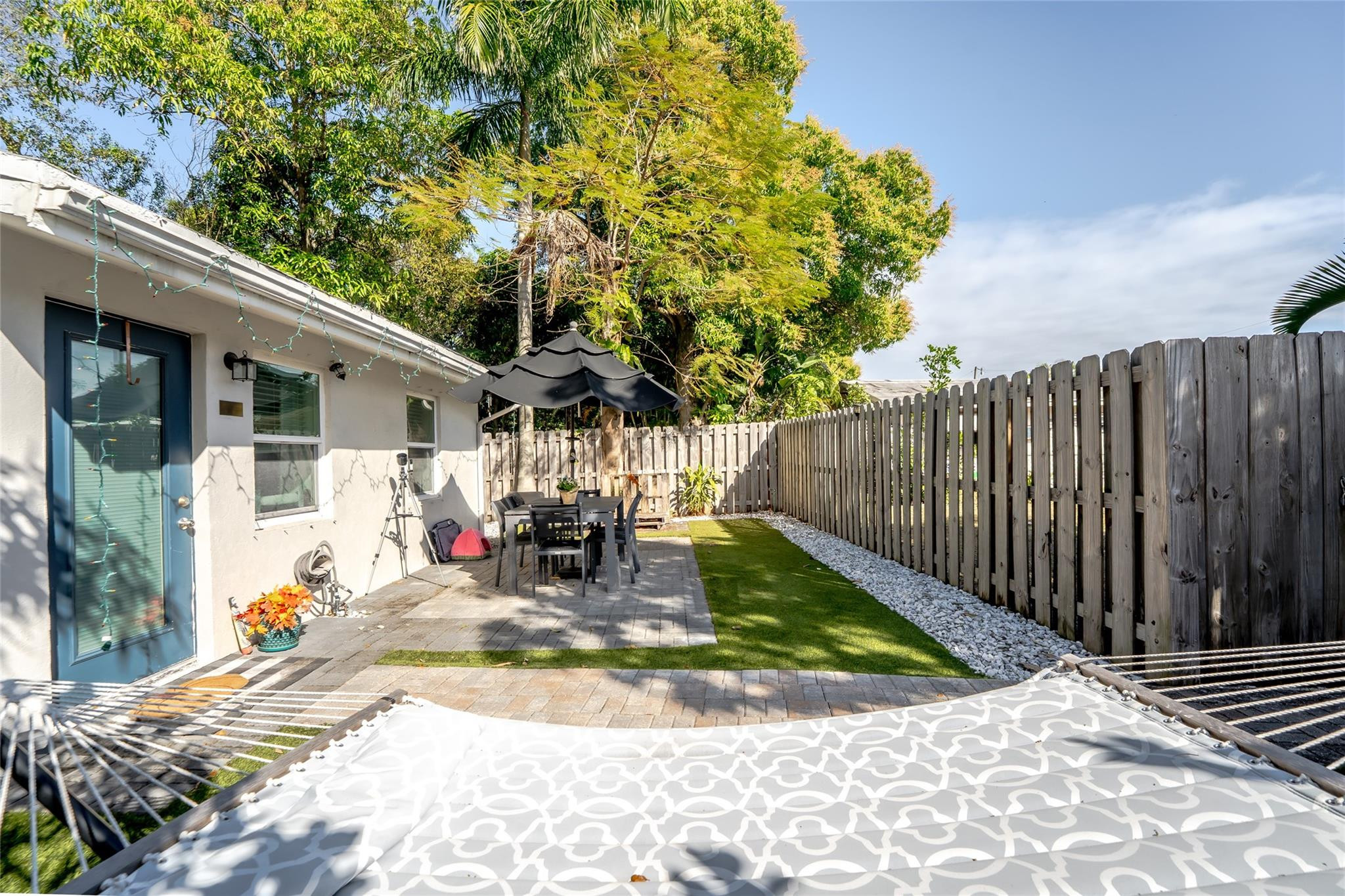 Fenced backyard patio with a paved dining area, umbrella, narrow lawn, and hammock.