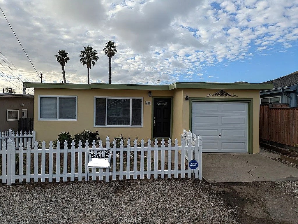 Single-story beige house with green trim, white picket fence, and a FOR SALE sign.