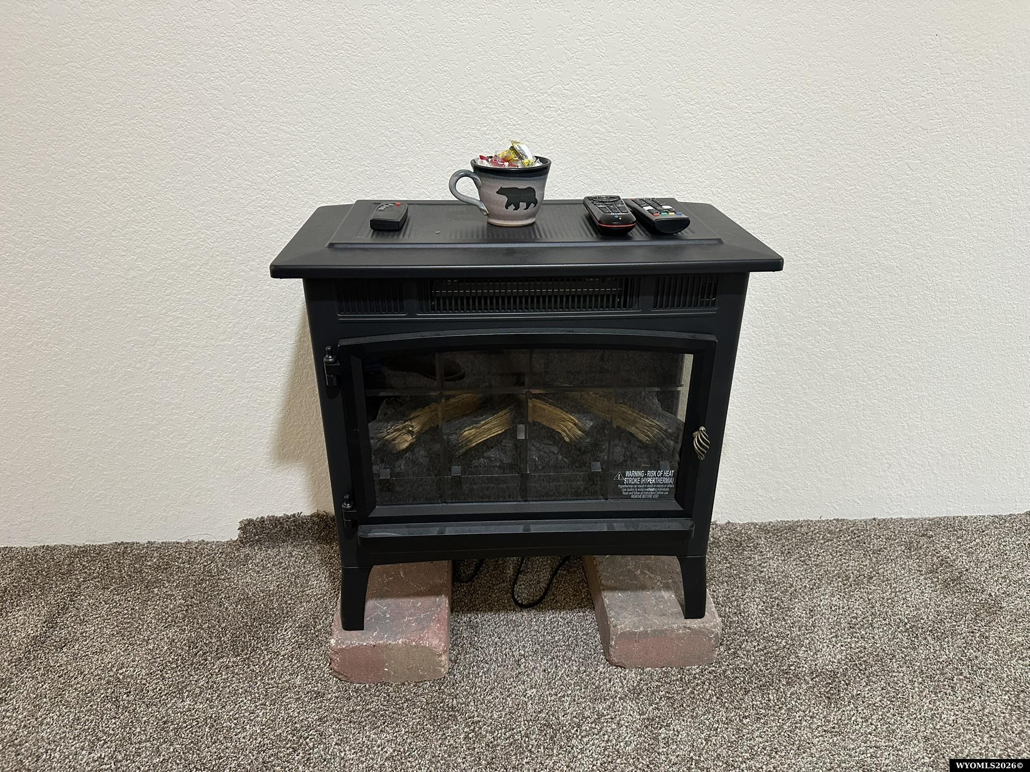 Black electric fireplace heater with faux logs, raised on bricks in a carpeted room.