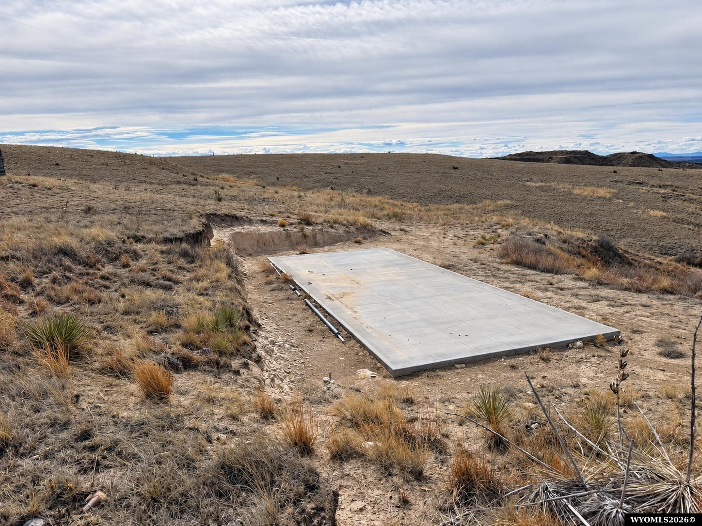 Vacant arid lot with a rectangular concrete pad and low rocky hills.