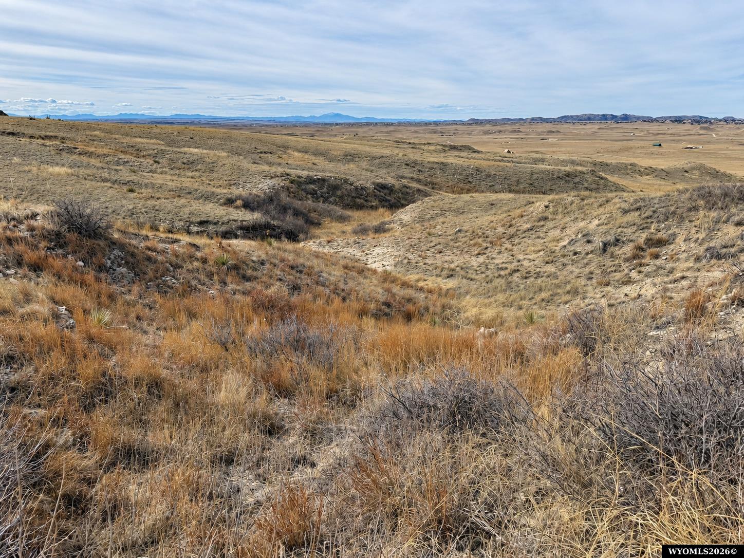 Rolling grassland lot with low brush and distant mountain ridges beneath a cloudy sky.
