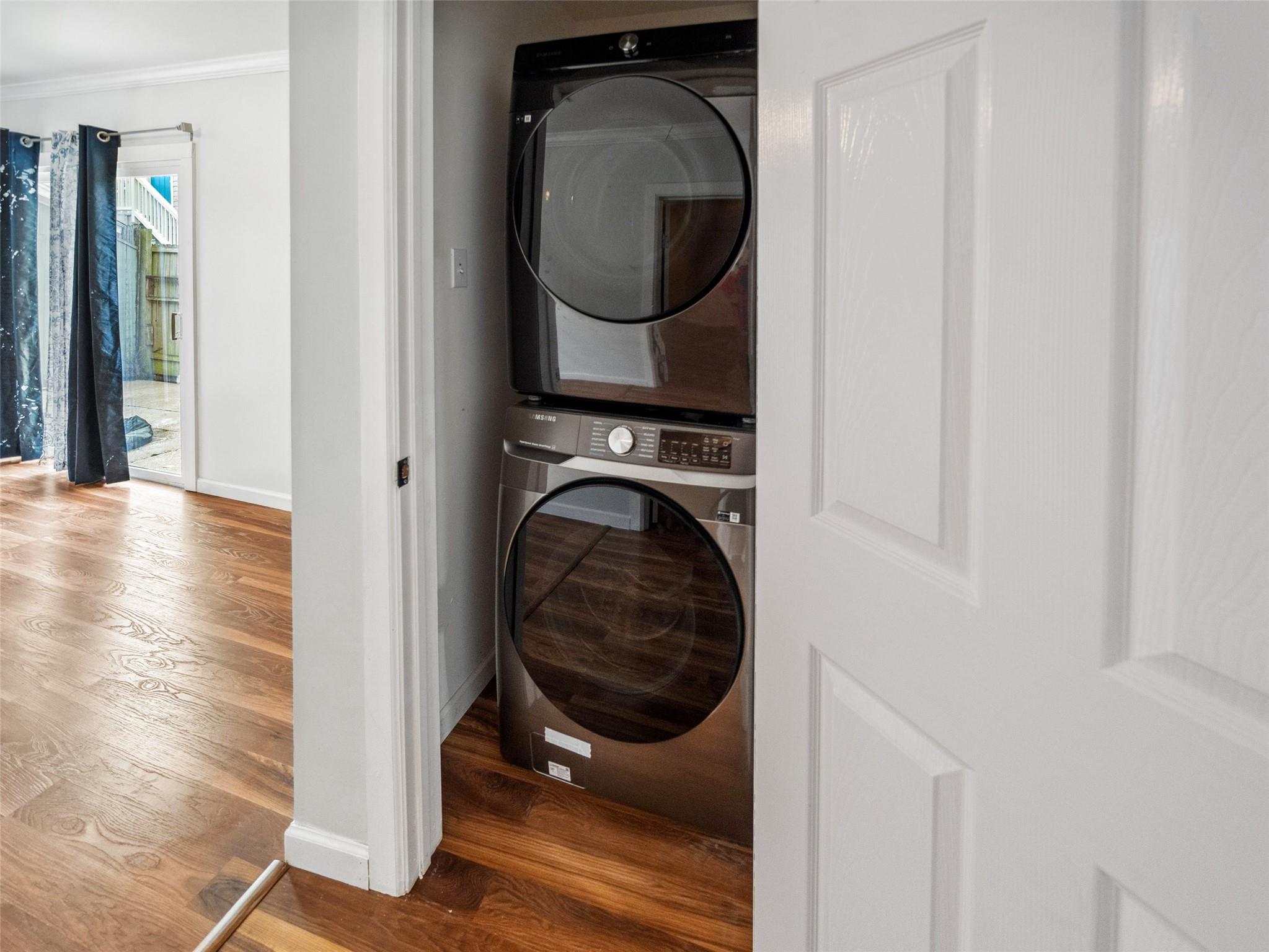 Stacked washer and dryer in a closet beside a hardwood-floored room with curtained patio doors.