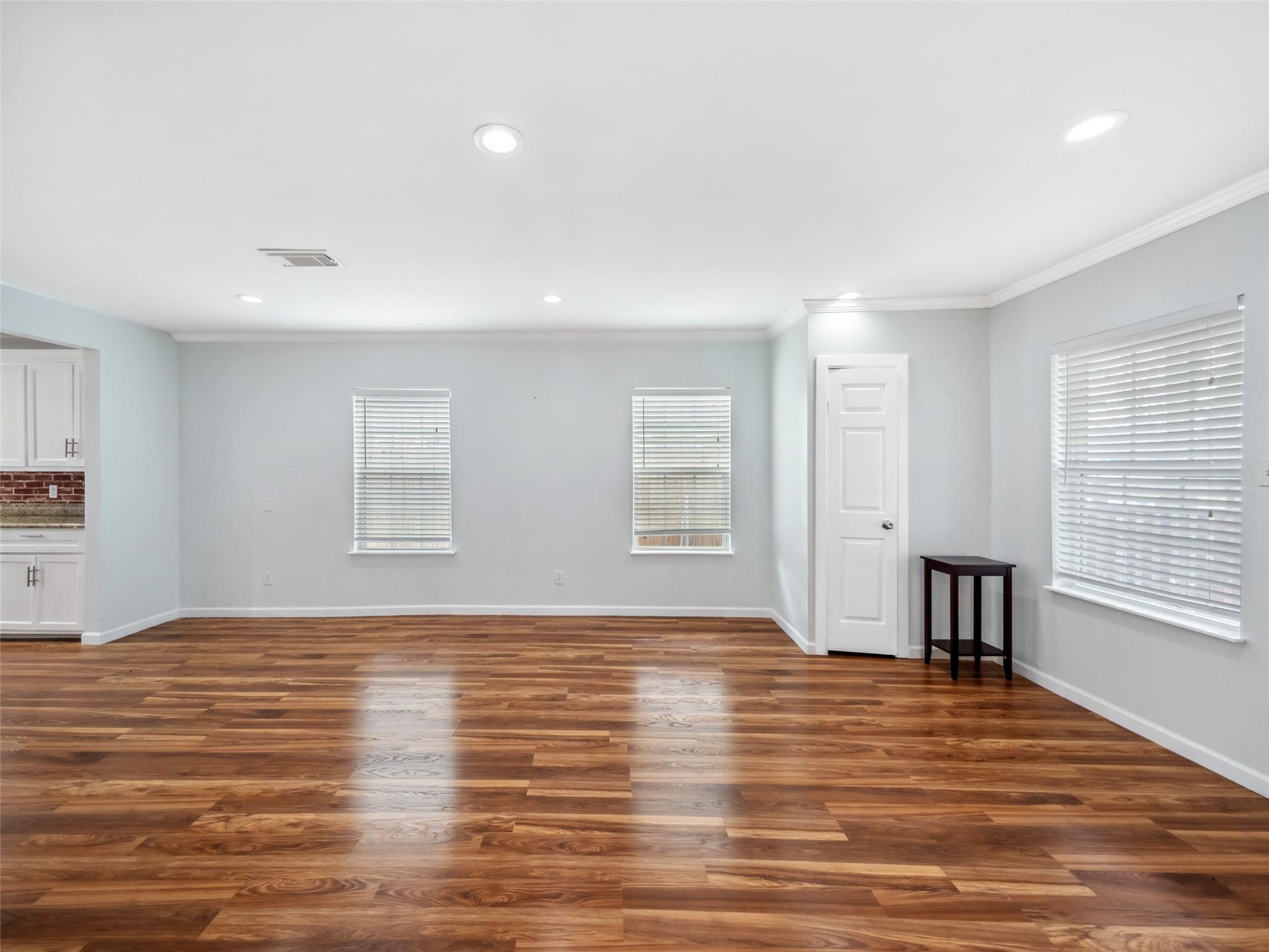 Mostly empty living room with glossy hardwood floors, three windows with blinds, and a small side table.