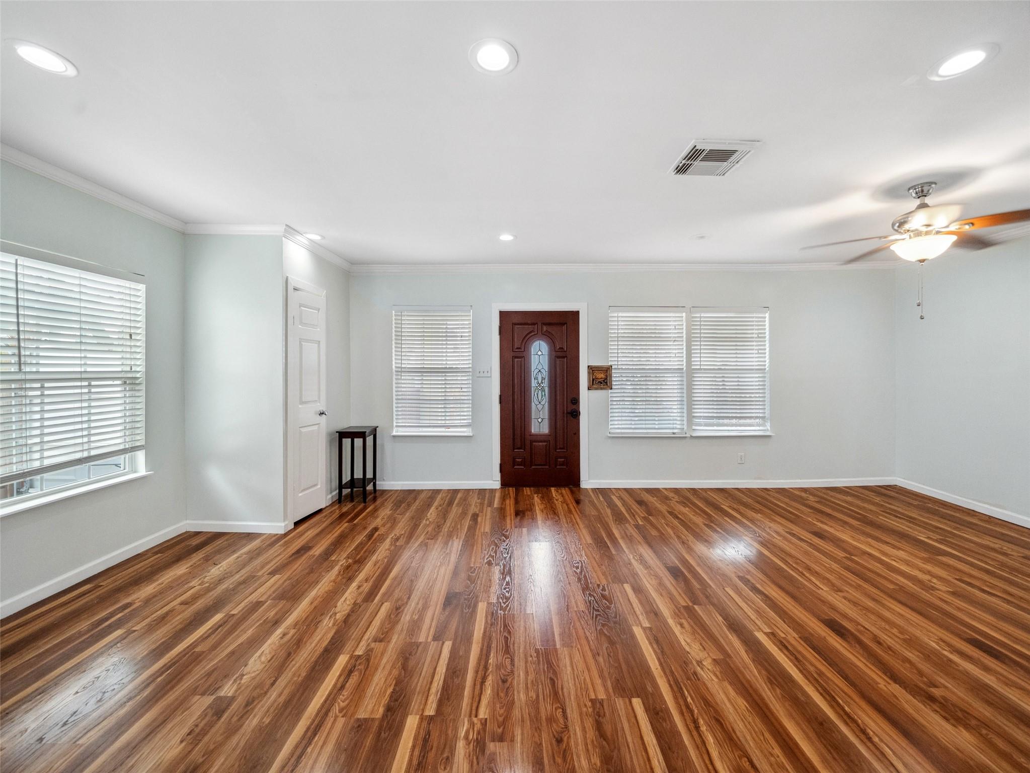 Empty living room with glossy wood floors, a dark front door, and windows with white blinds.