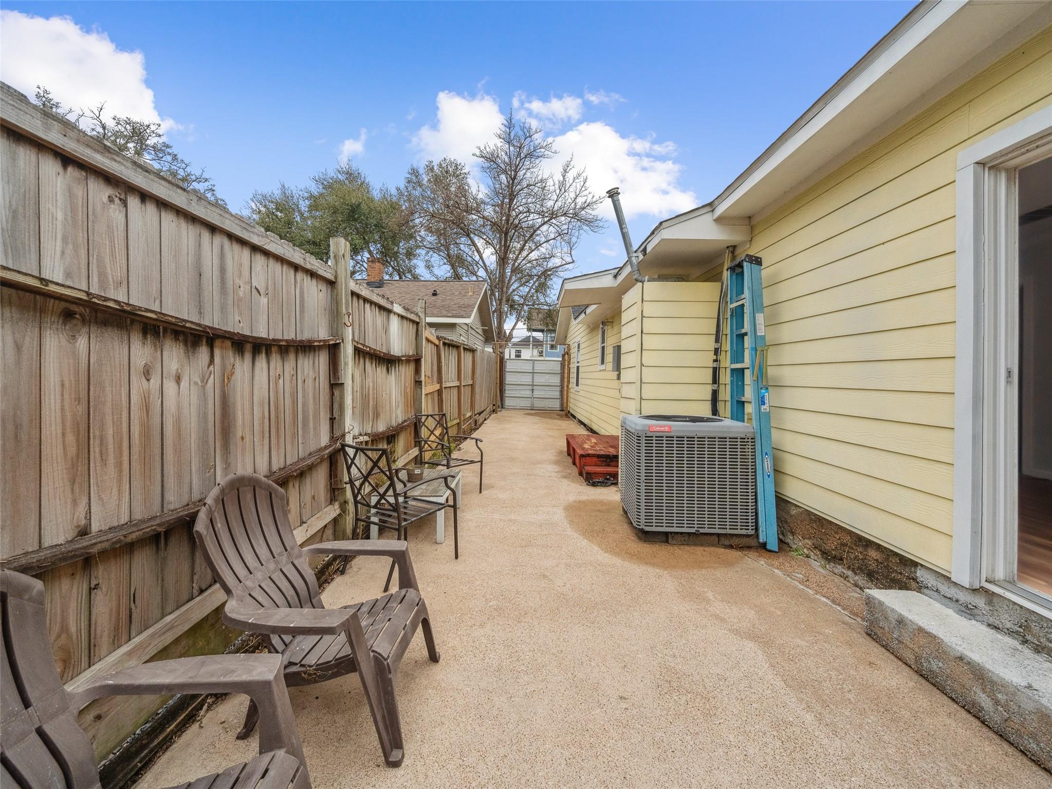 Concrete side patio with wooden fence, outdoor chairs, and an air-conditioning unit beside a yellow house.