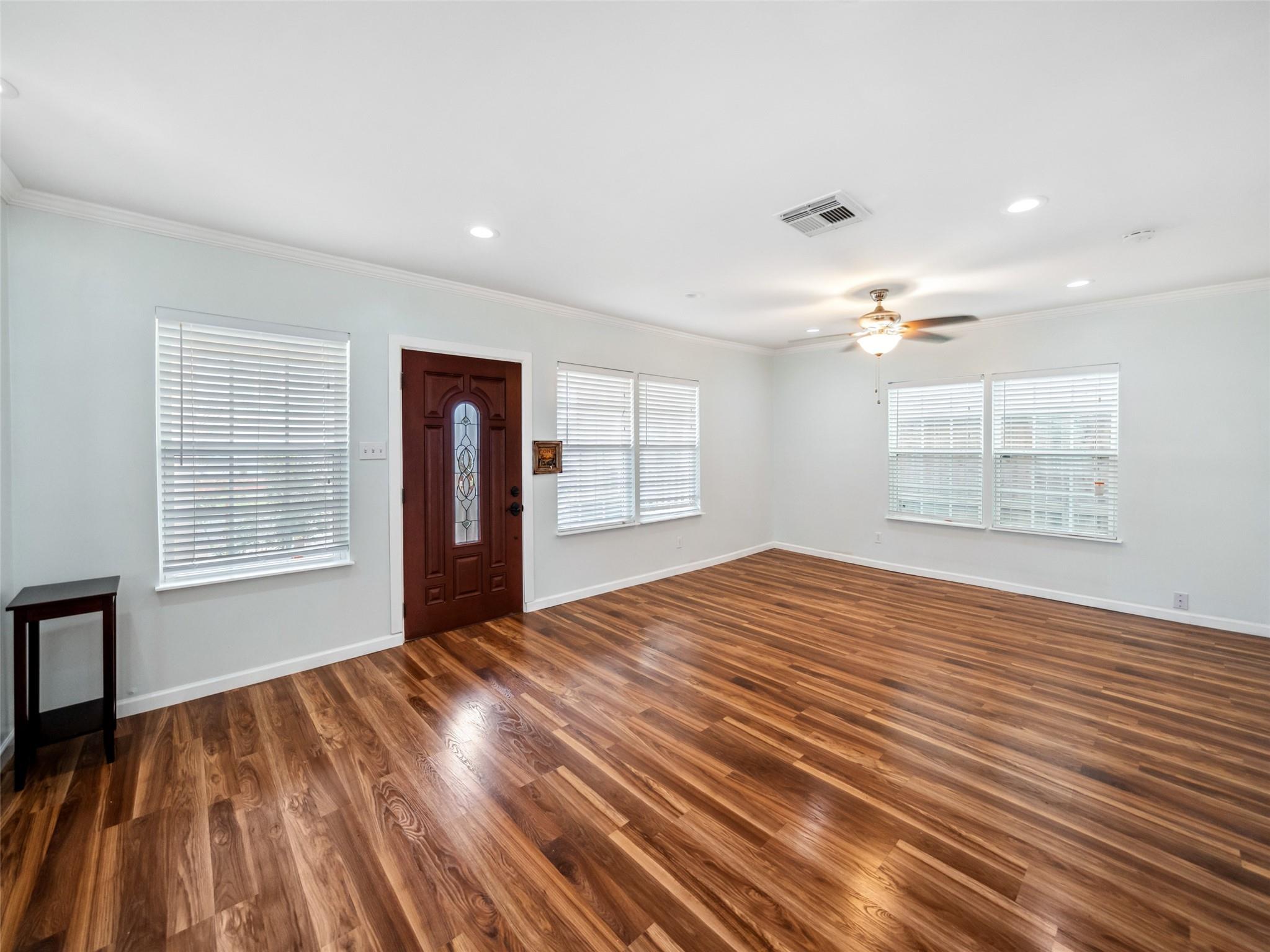 Empty living room with glossy wood floors, multiple blinded windows, a decorative front door, and ceiling fan.